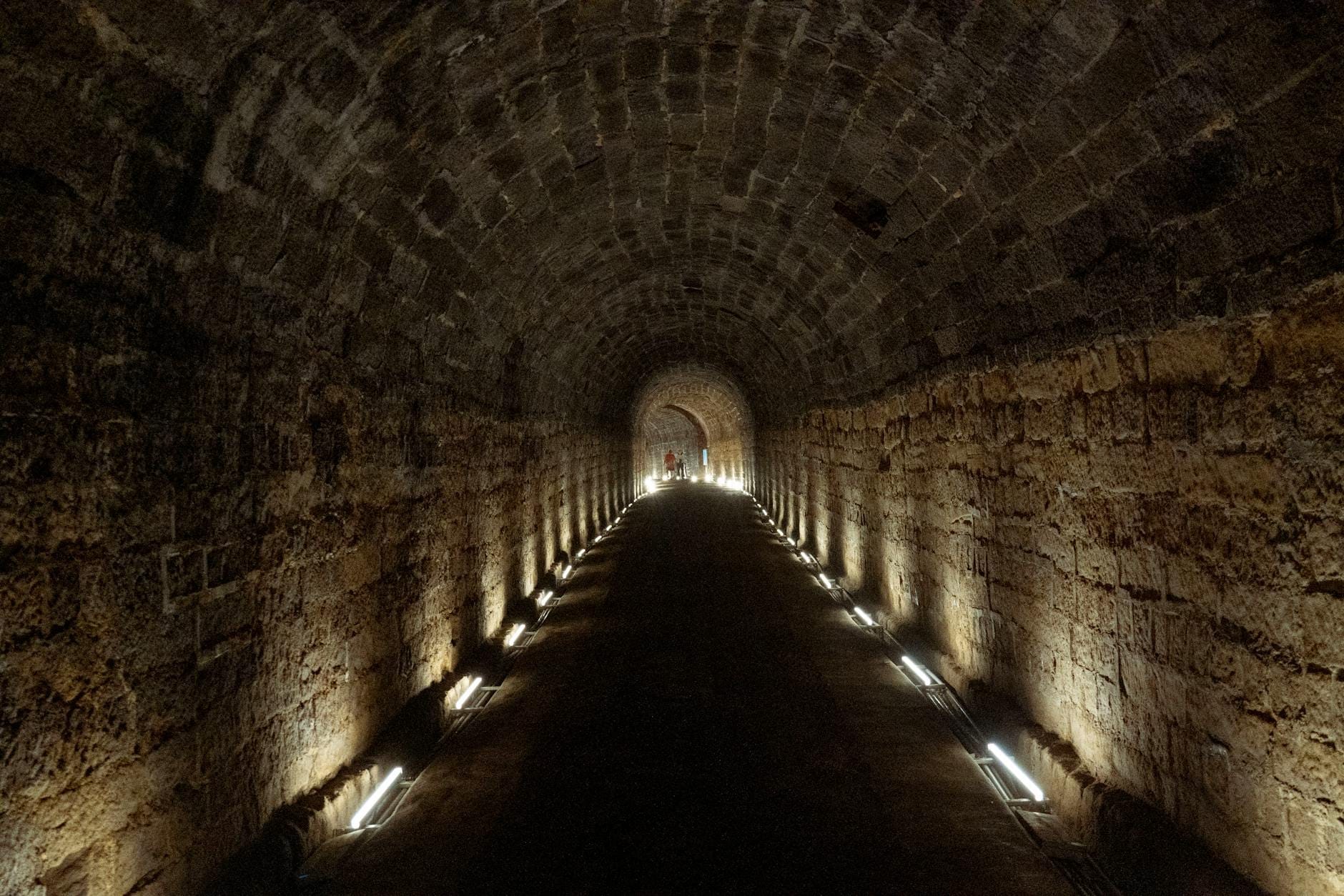 Túnel subterrâneo de pedra bem iluminado com teto abobadado e corredor, com luzes ao longo de ambas as paredes, visto de frente.
