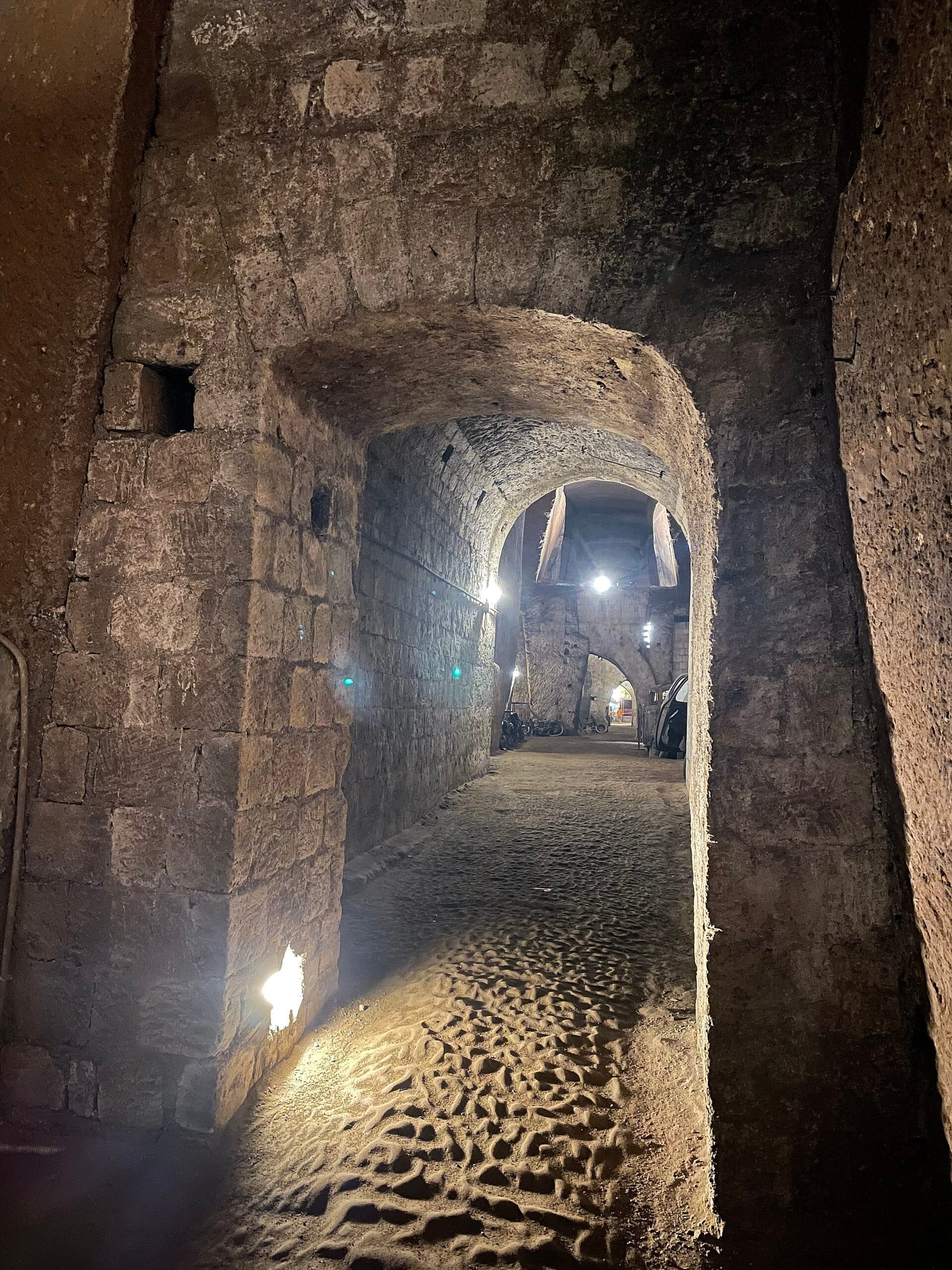 Archi in pietra e pareti grezze dei tunnel della Napoli Sotterranea, illuminati da una luce soffusa, che mostrano l'antica architettura sotterranea sotto Napoli.