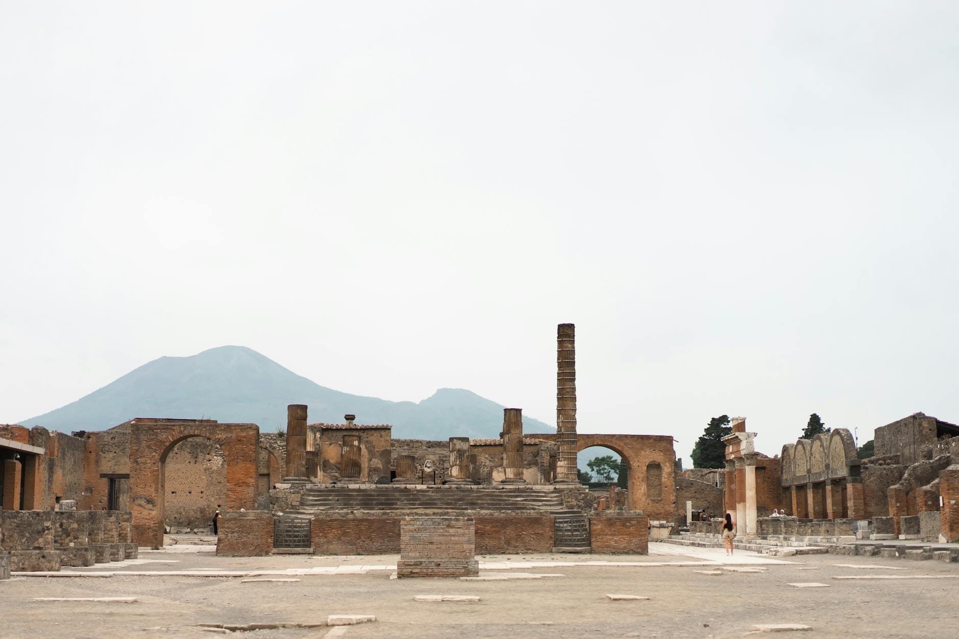 Vista ampla das ruínas de Pompeia com o Monte Vesúvio ao fundo sob um céu pálido, mostrando antigas estruturas de pedra e espaços abertos.
