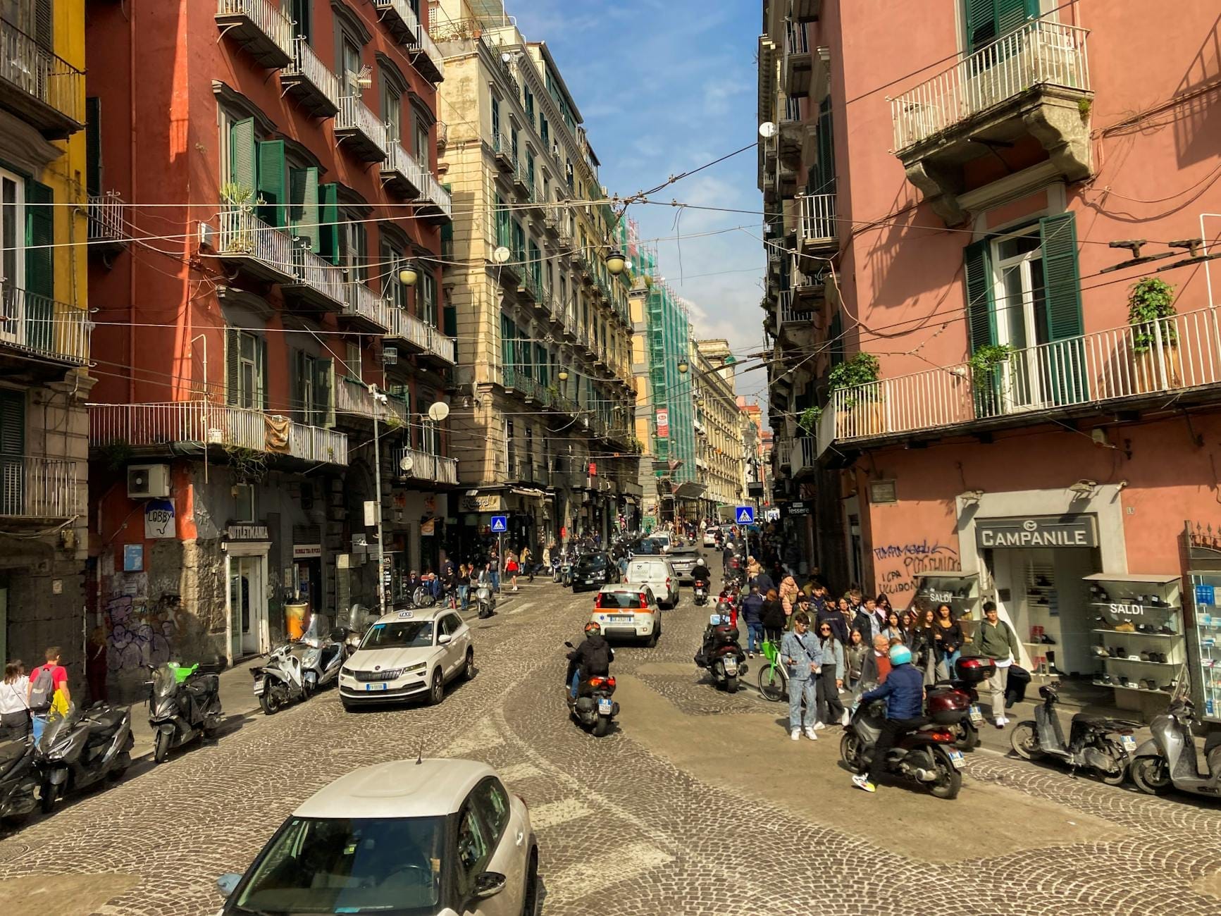 Scène de rue animée à Naples avec des passants, des scooters, des cafés et de colorés bâtiments historiques longeant une rue pavée.