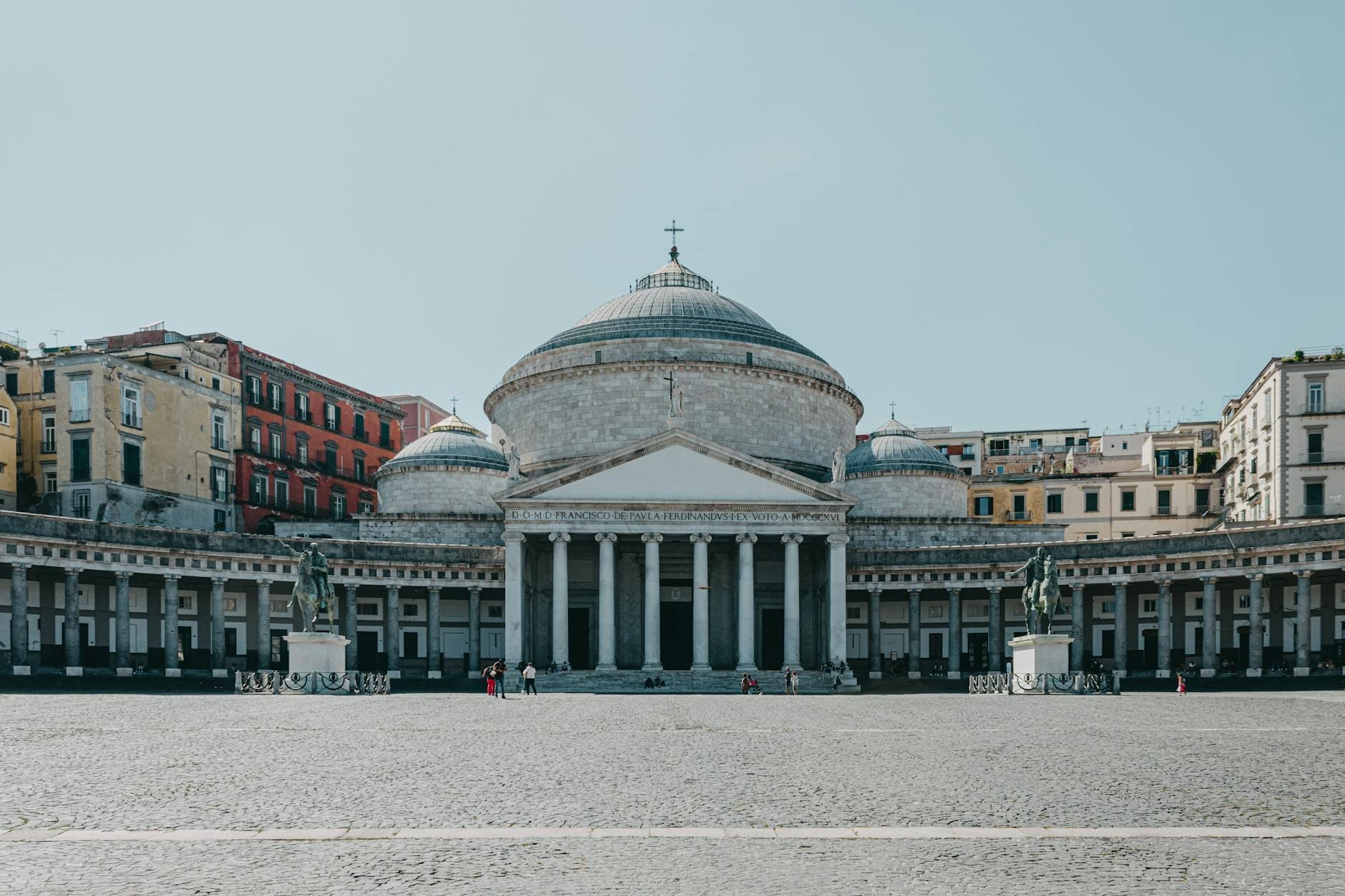 Vue large et dégagée de la Piazza del Plebiscito à Naples, montrant la grande église à coupole et les bâtiments environnants sous un ciel bleu.