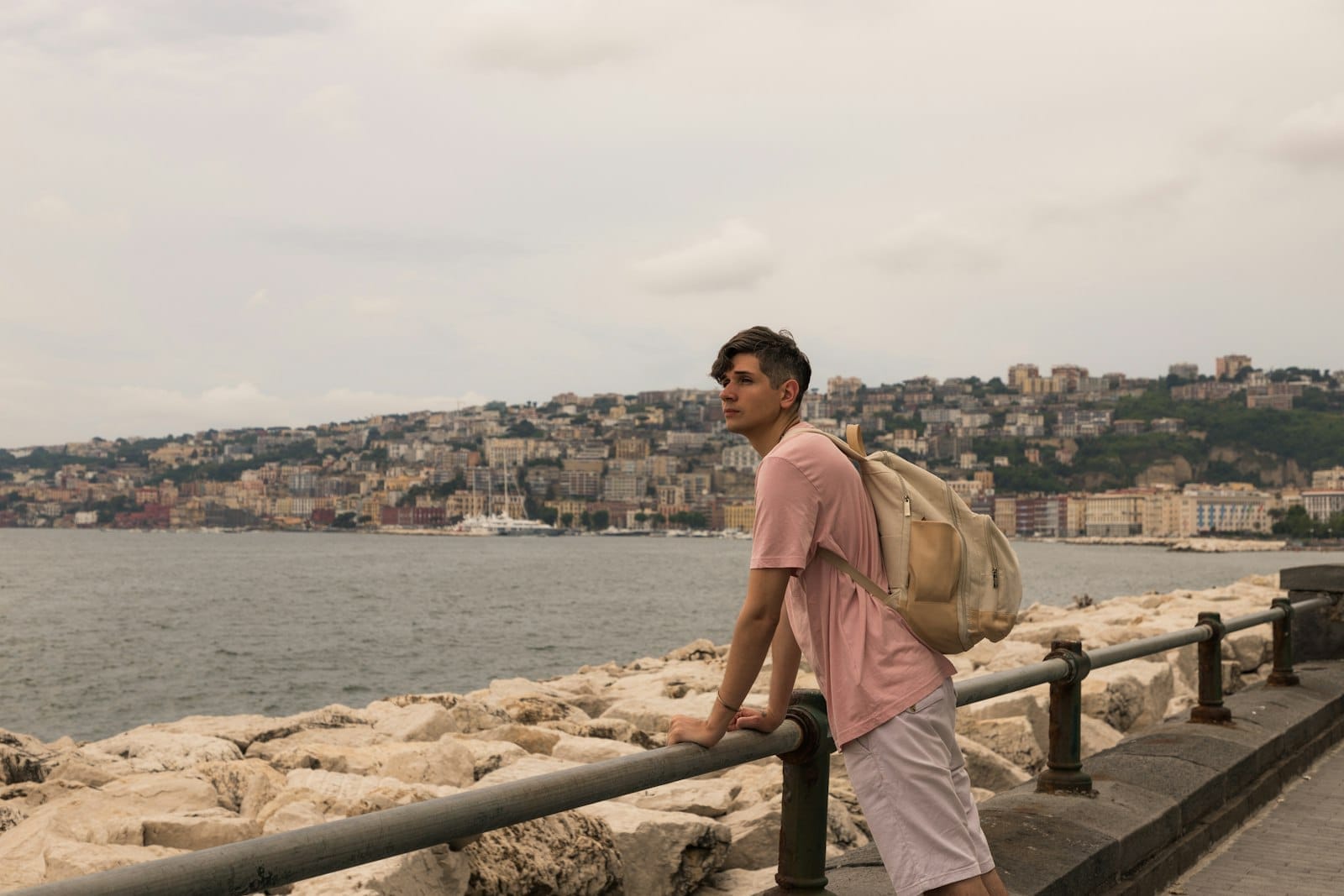 Um jovem com uma mochila se apoia em um corrimão à beira-mar em Nápoles, com edifícios da cidade e colinas escuras ao fundo.