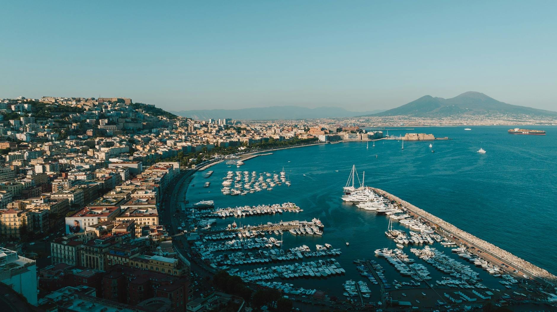 A wide panoramic view of Naples’ bay with yachts in the marina, city buildings, and Mount Vesuvius in the background under a clear blue sky.