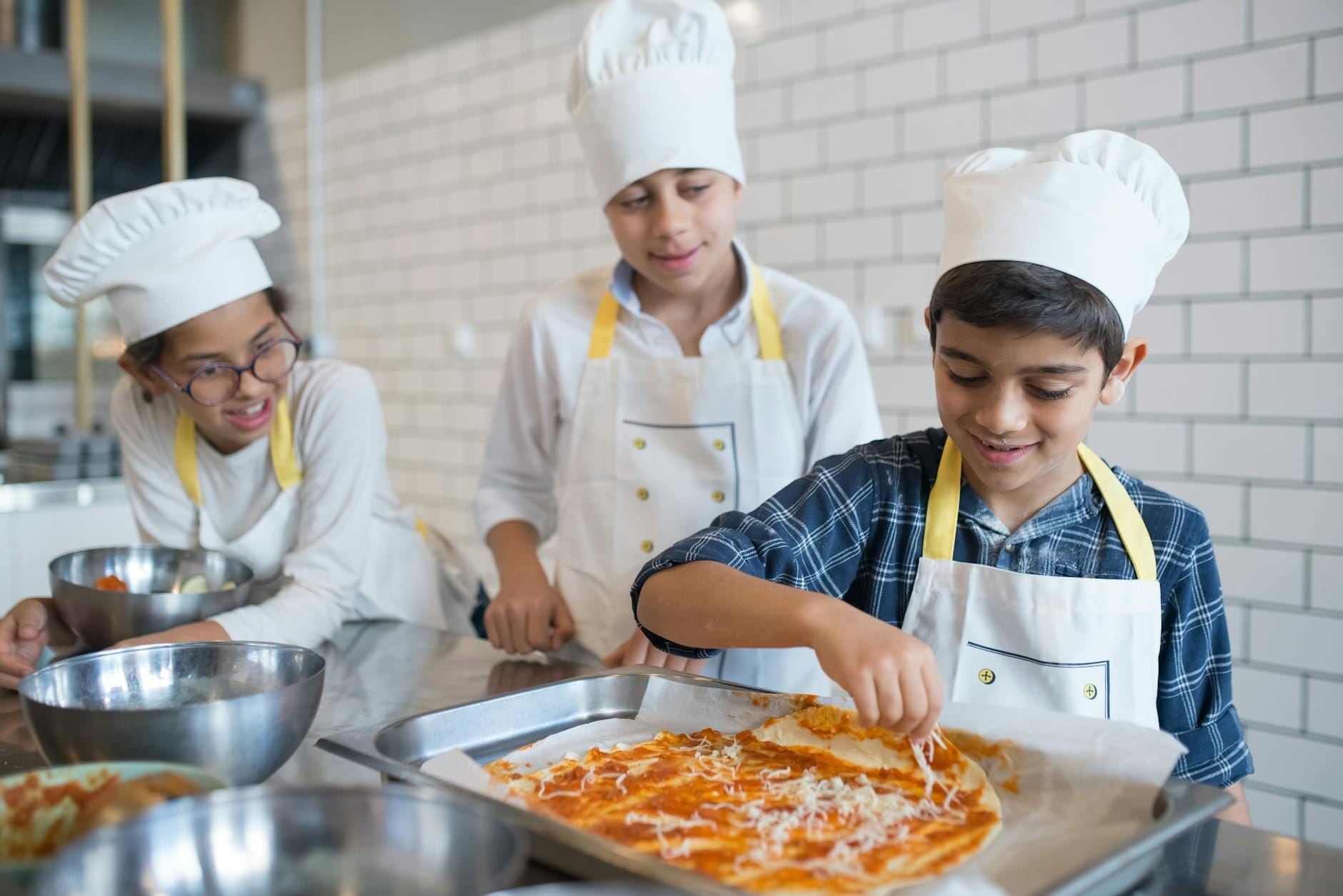 Três crianças com chapéus e aventais de chef fazendo pizza juntas em uma assadeira em uma cozinha moderna com paredes de azulejos brancos.