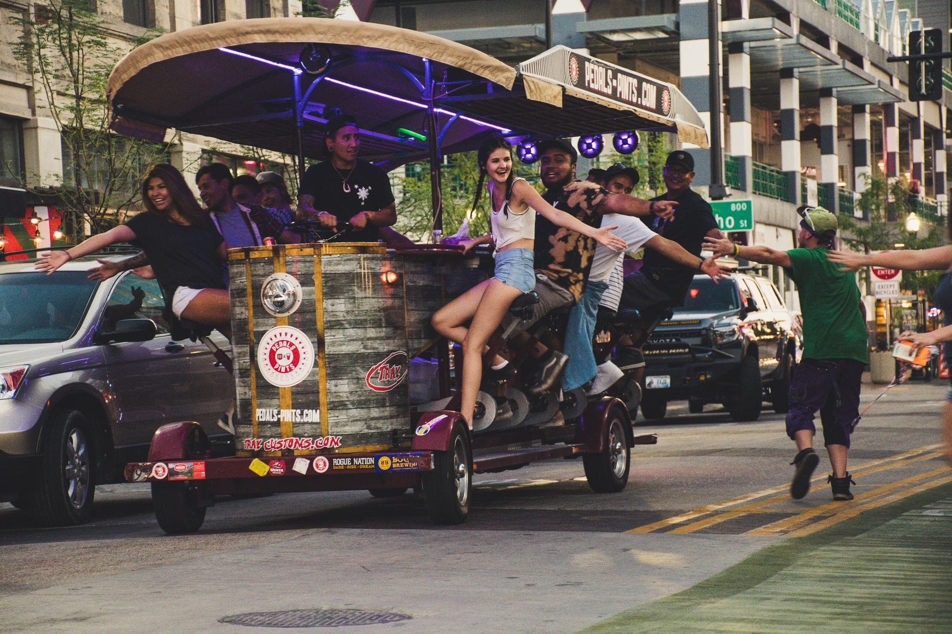 Un animado grupo de personas disfrutando de un paseo en taberna de pedales con luces de neón, moviéndose por una concurrida calle en un entorno urbano.