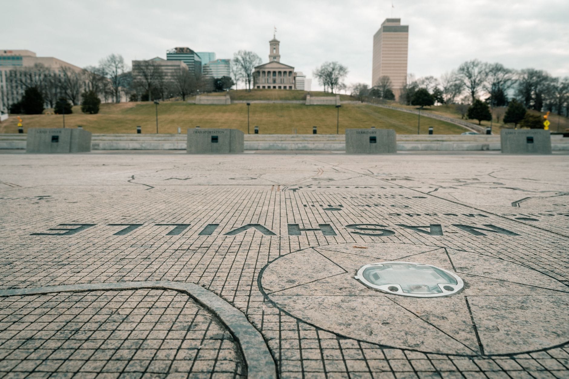 Vista del Capitolio del Estado de Tennessee en una colina, visto desde el Parque del Bicentenario con un mapa de piedra de Nashville en primer plano bajo un cielo nublado.