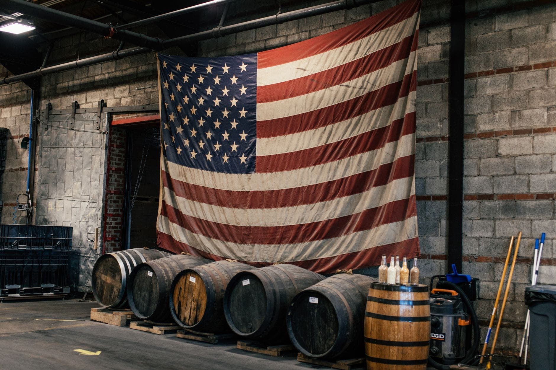 Fila de barriles de whiskey en el interior de una destilería urbana con una gran bandera americana colgada en una pared de ladrillo.