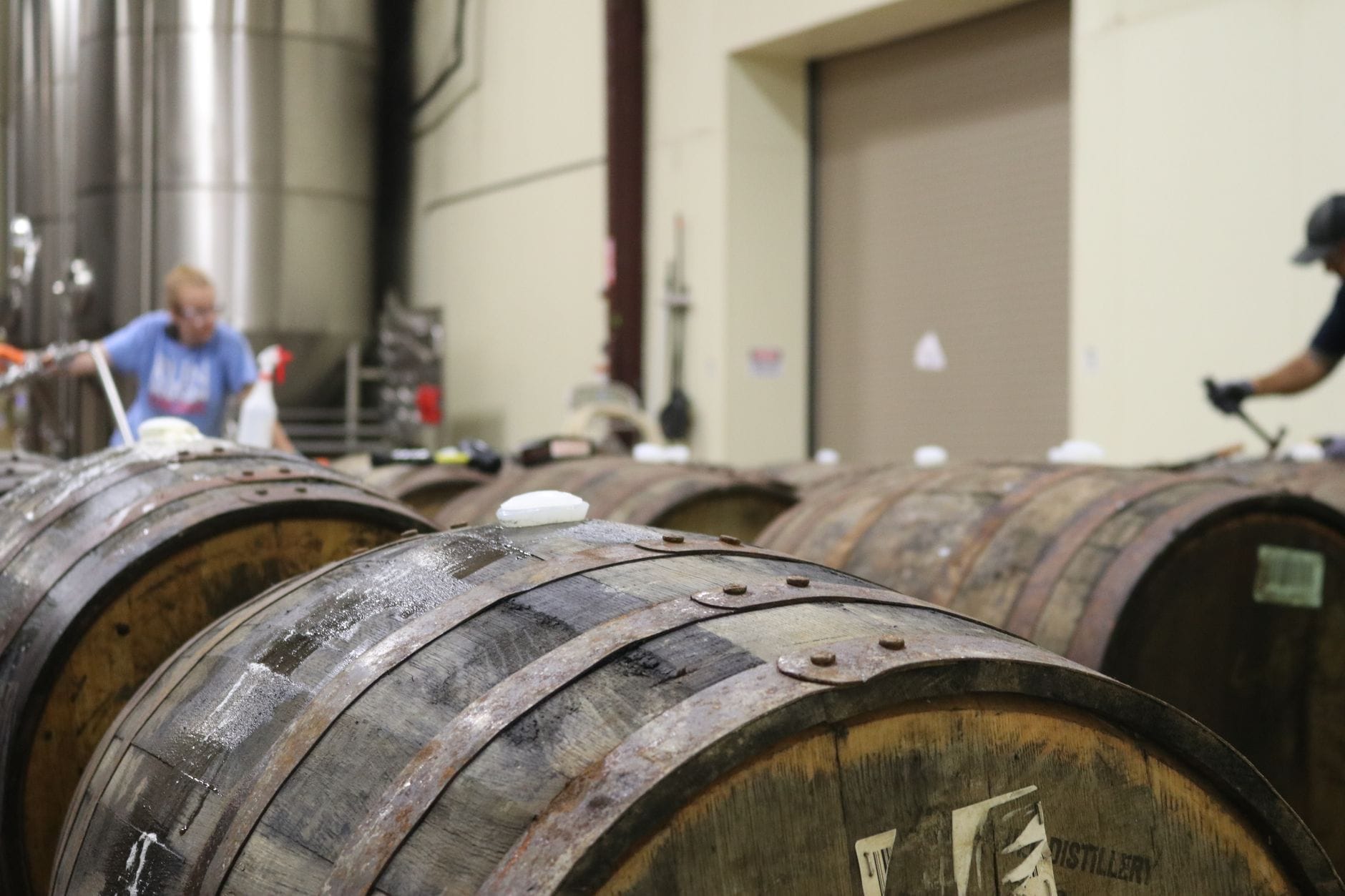 Hileras de barriles de whiskey en el interior de un almacén de destilería, con trabajadores al fondo atendiendo los barriles.
