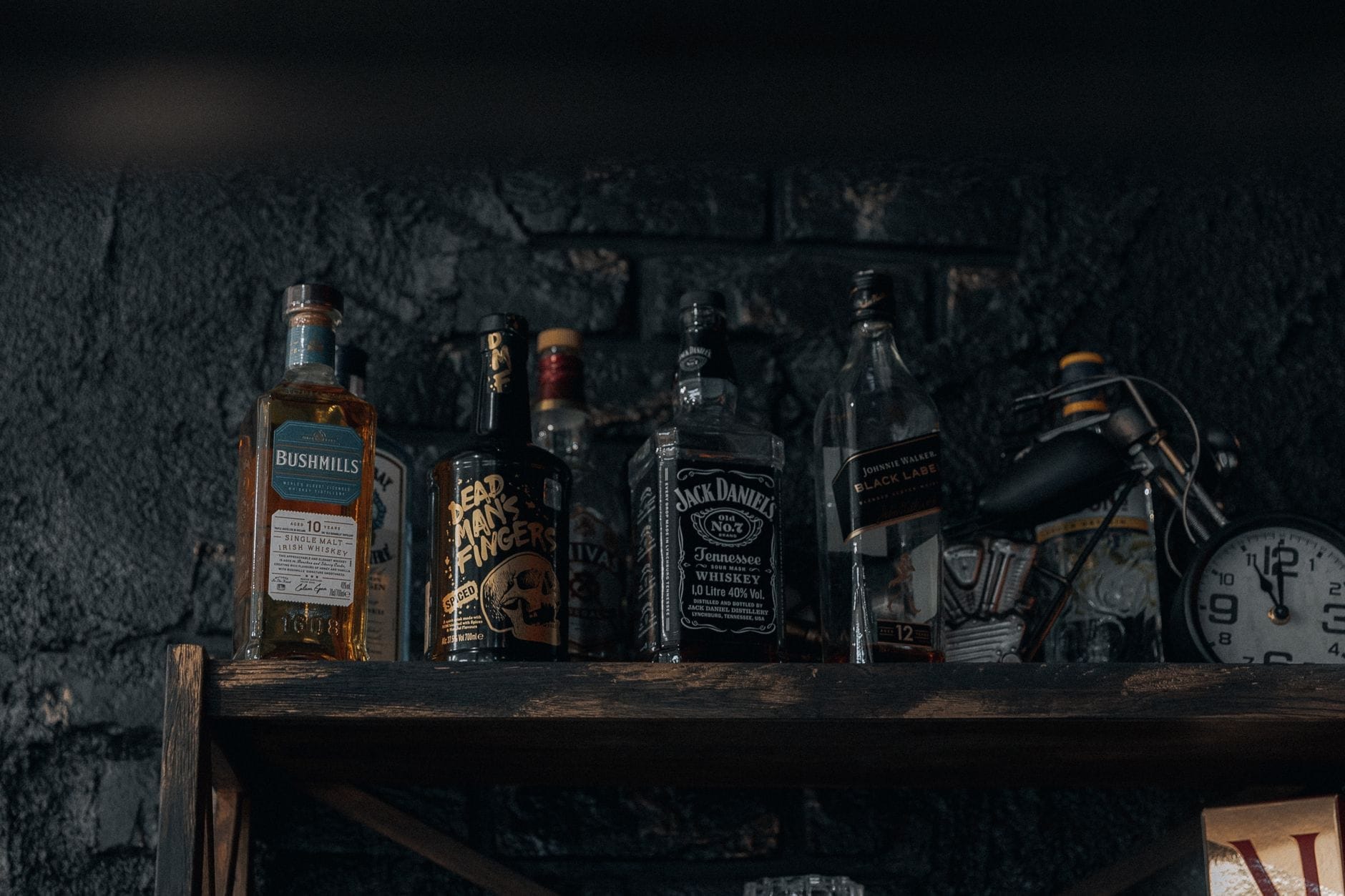 Un estante de botellas de whiskey incluyendo Jack Daniel's sobre un fondo oscuro y rústico, con un reloj y pequeños objetos decorativos cerca.