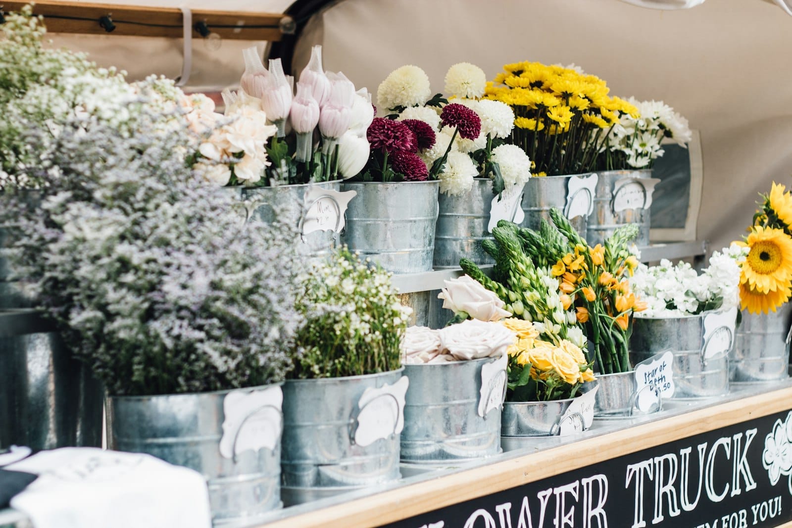 Cubos con flores frescas de colores variados sobre un puesto de un mercado agrícola al aire libre, con un ambiente vibrante y acogedor.