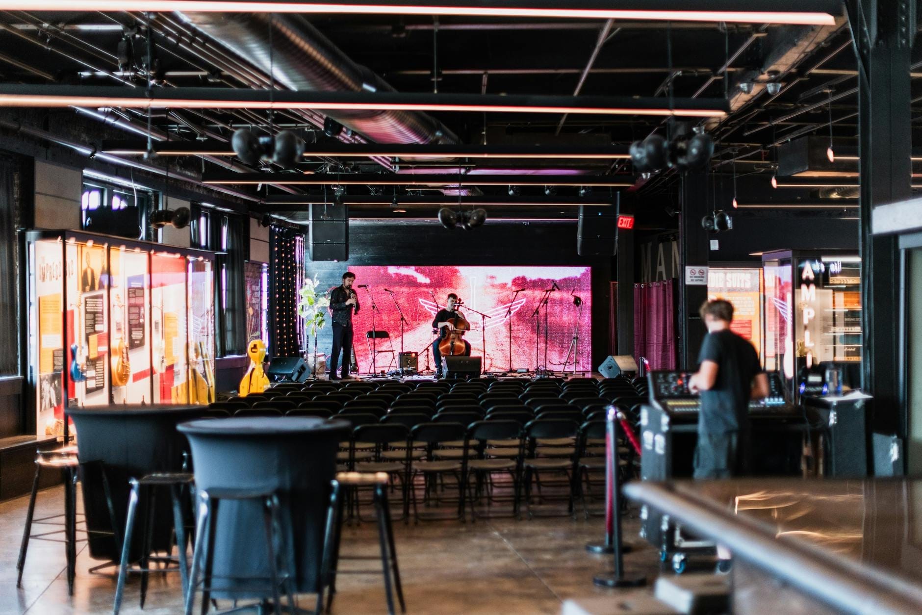 Sala de música en vivo moderna con un pequeño escenario, sillas vacías y algunos músicos preparándose, probablemente en un barrio de Nashville.