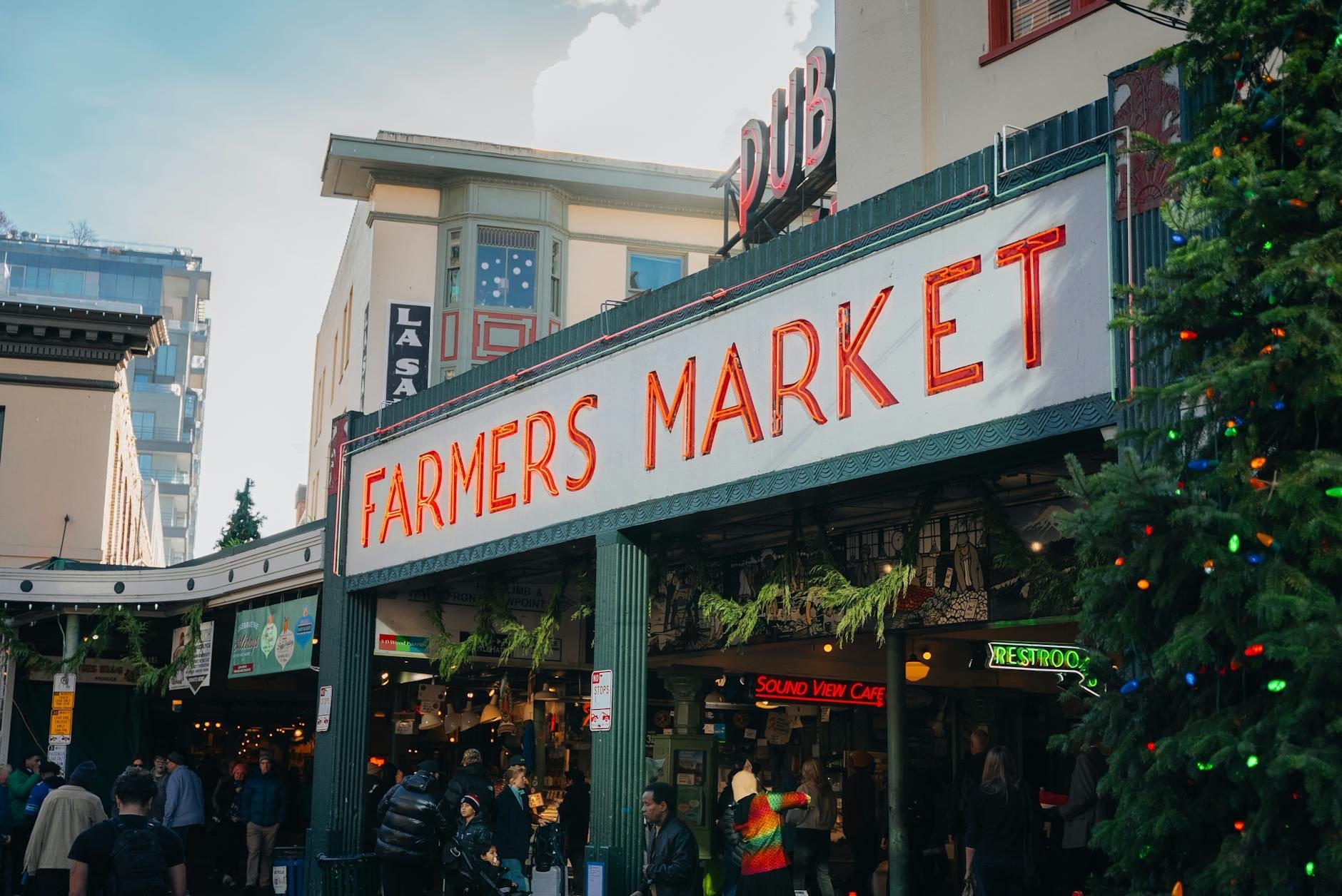 Entrada a un animado mercado de agricultores decorado para las fiestas, con personas comprando y un árbol de Navidad visible.