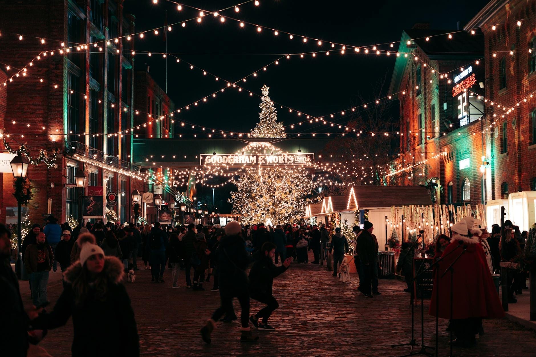 Mercado navideño al aire libre de noche con personas caminando bajo guirnaldas de luces, un gran árbol de Navidad iluminado y edificios históricos de ladrillo decorados para las fiestas.