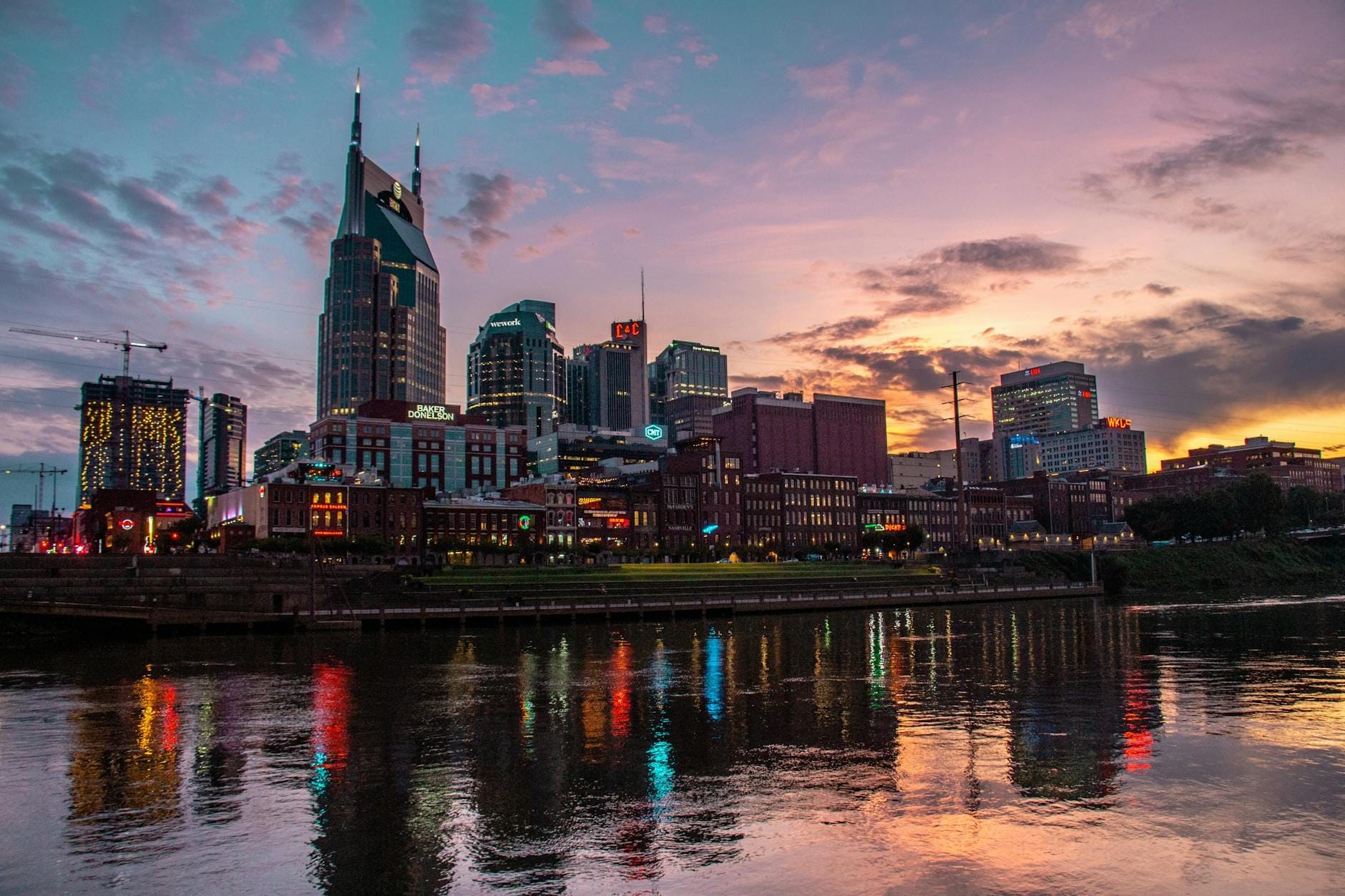 Panorámica del centro de Nashville al atardecer con edificios de lujo y luces de colores reflejadas en el río Cumberland, evocando una atmósfera vibrante y sofisticada.