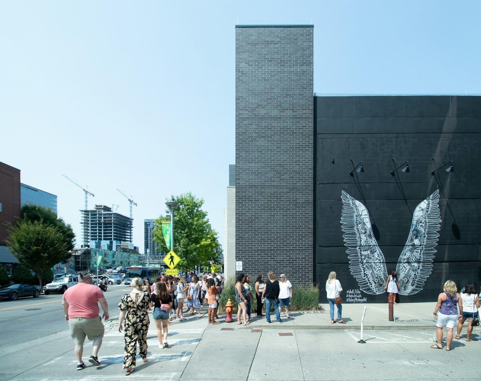 Vista amplia de la calle con el icónico mural negro de alas blancas en Nashville, con personas haciendo fila para tomarse fotos y edificios de la ciudad al fondo.