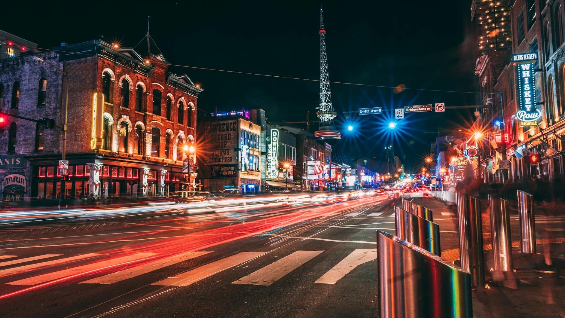 Animada escena nocturna de Nashville con letreros de neón, edificios históricos de ladrillo y estelas de luces de autos que capturan la energía de la vida nocturna de la ciudad.