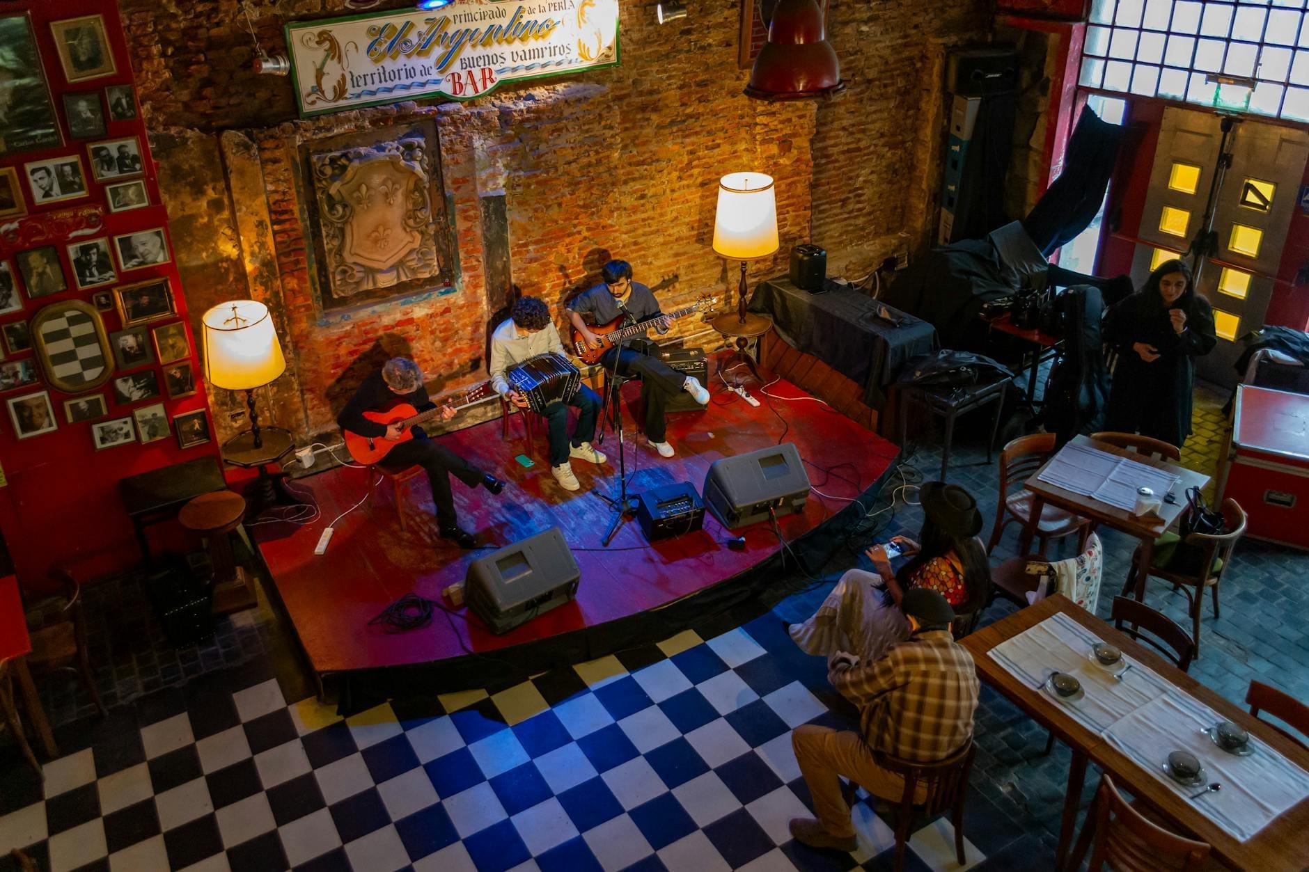 Vista aérea de músicos actuando en un pequeño escenario en una acogedora sala de escucha con paredes de ladrillo y algunas personas sentadas en mesas.