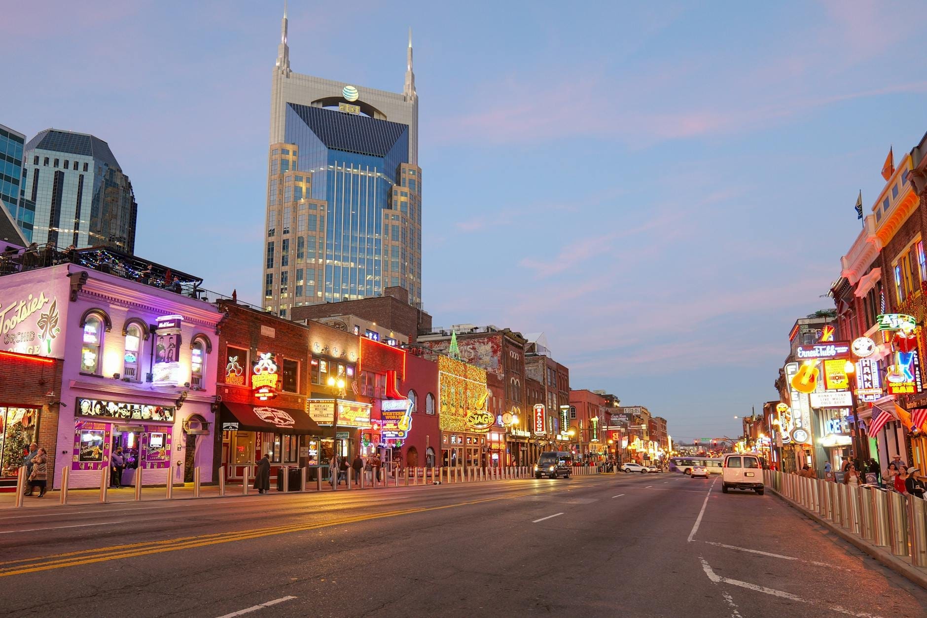 Vista vibrante del centro de Nashville al atardecer: Broadway iluminado con letreros de neón, locales de música en vivo y el edificio AT&T bajo un cielo rosa y azul.