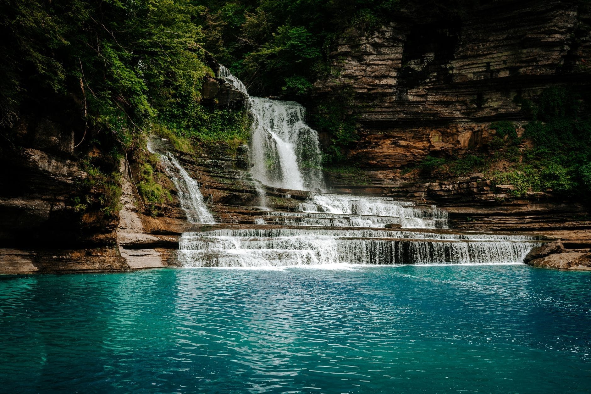 Amplia cascada escalonada con poza de agua turquesa rodeada de acantilados rocosos y exuberantes árboles verdes en un día soleado.