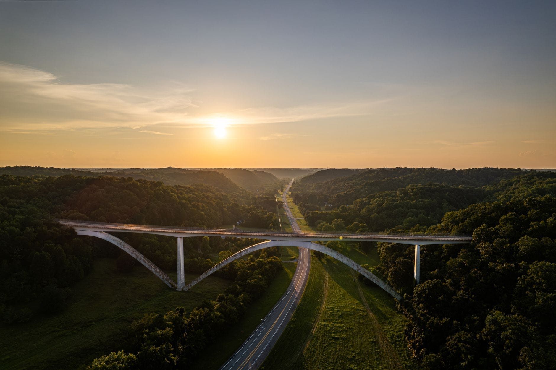 Vista aérea panorámica de un puente sobre una carretera escénica rodeada de colinas verdes al atardecer, evocando el inicio de un hermoso road trip por Tennessee.