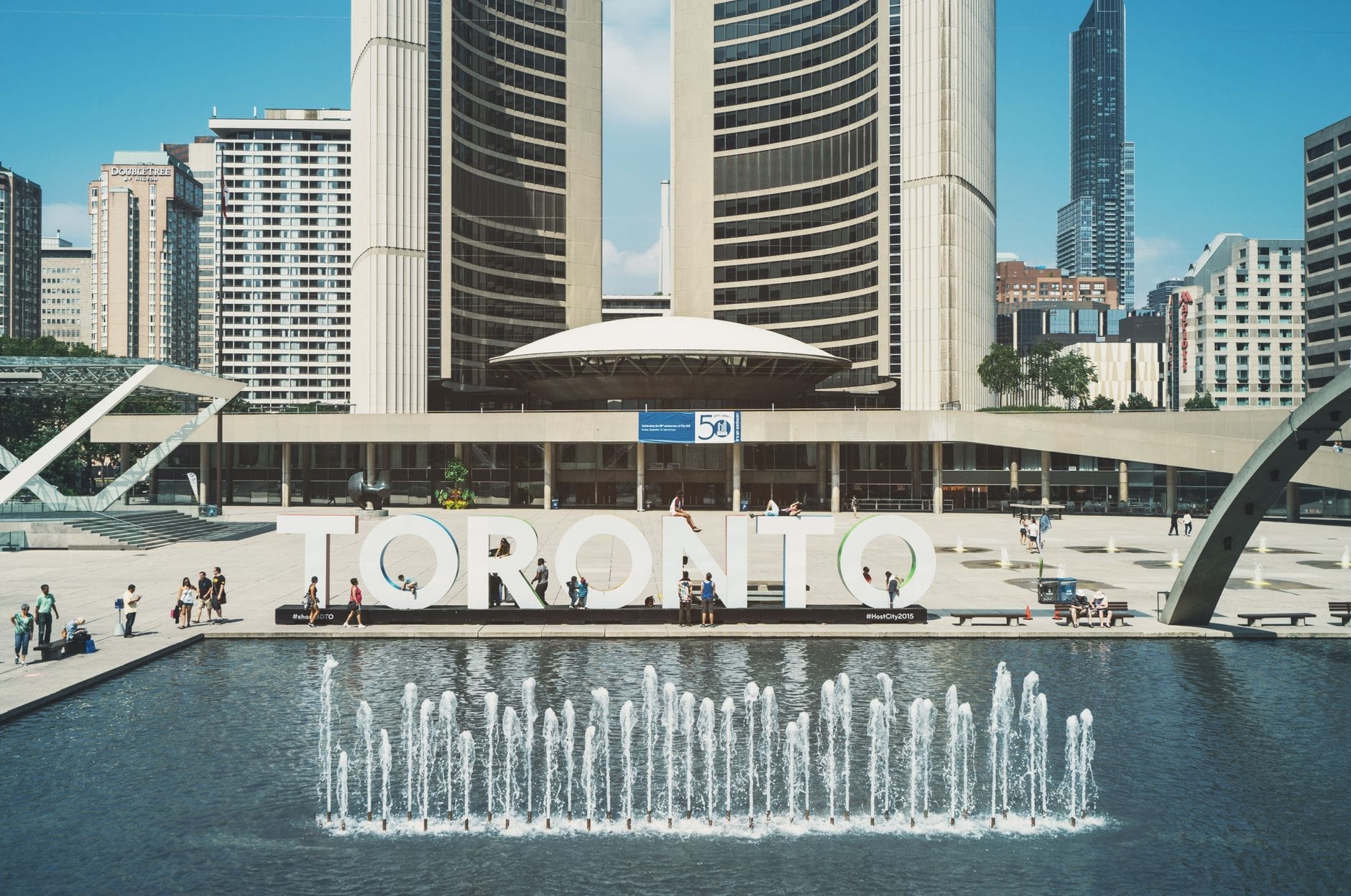 สนามนาธาน ฟิลลิปส์ยามกลางวัน มีศาลากลางเมืองโตรอนโต ป้าย TORONTO น้ำพุ และผู้คนเดินเล่นหน้าตึกสไตล์โมเดิร์น