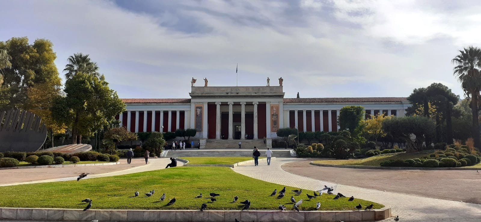 Vista frontale del National Archaeological Museum Athens con colonne neoclassiche, statue sul tetto, giardini verdi e persone che camminano nelle vicinanze.