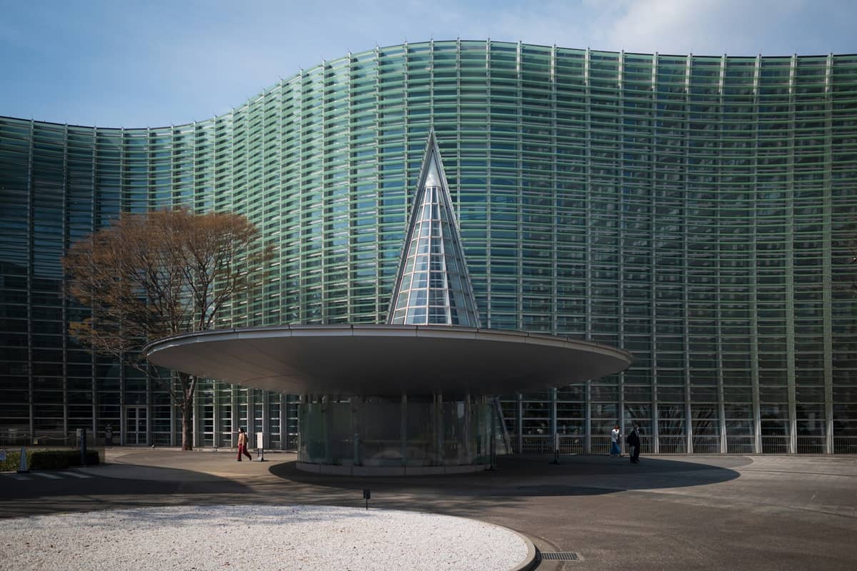 Exterior of The National Art Center, Tokyo featuring its distinctive wavy glass facade and unique circular entrance structure under blue sky.