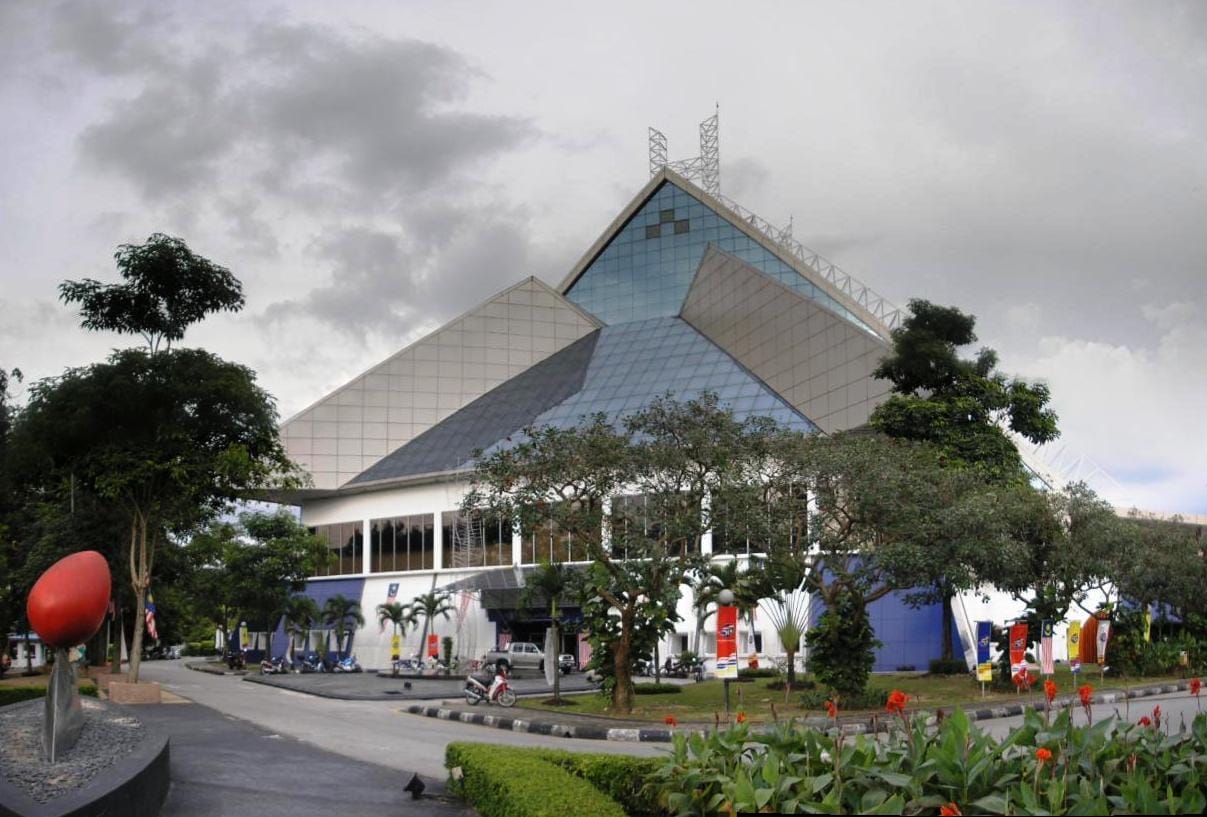 Exterior view of the National Art Gallery Kuala Lumpur, featuring its geometric glass and steel architecture, lush greenery, and colorful sculpture under cloudy skies.