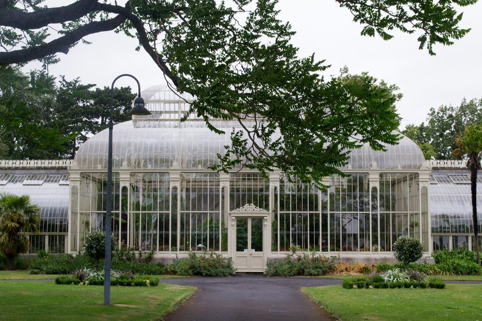 Serra vittoriana restaurata ai Giardini Botanici Nazionali di Glasnevin, circondata da rigogliosa vegetazione e giardini, vista dal percorso principale in una giornata nuvolosa.