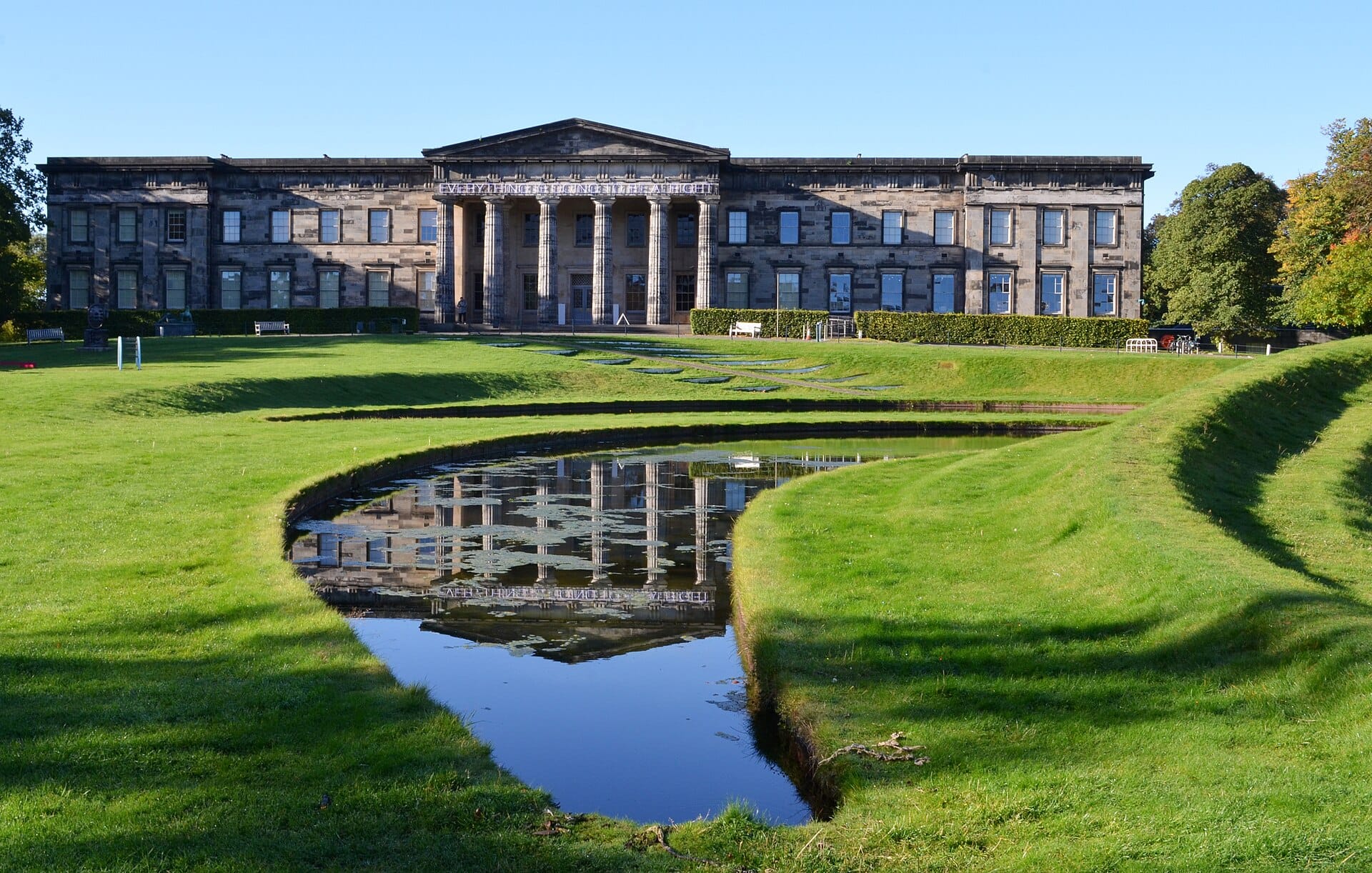 Die Scottish National Gallery of Modern Art (Modern One) mit Säulen, Skulpturen und dem markanten Teich mit Rasenflächen im Vordergrund an einem sonnigen, klaren Tag.