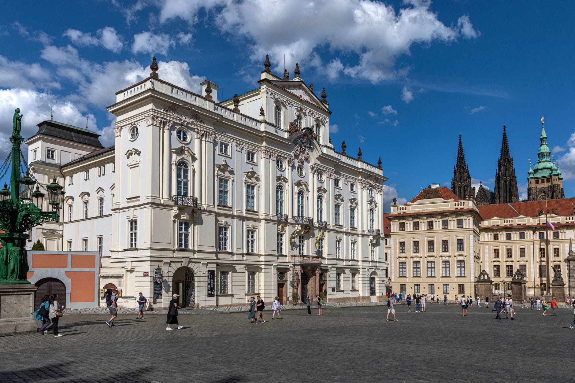 Weitwinkelperspektive auf die elegante Barockfassade des Sternberg-Palais am Hradschin-Platz mit Passanten und den Türmen der Prager Burg im Hintergrund unter strahlend blauem Himmel.