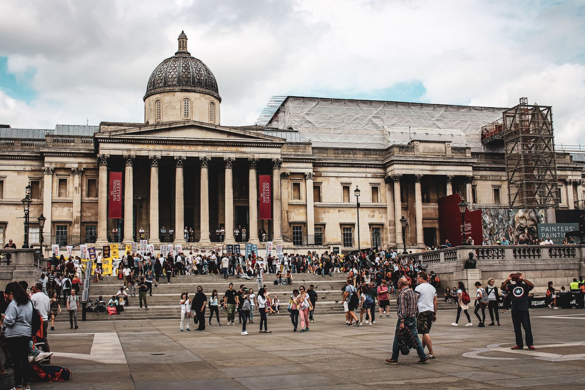 Vista ampia della National Gallery di Londra con la sua imponente facciata, la cupola centrale e la folla di visitatori fuori su Trafalgar Square sotto un cielo nuvoloso.