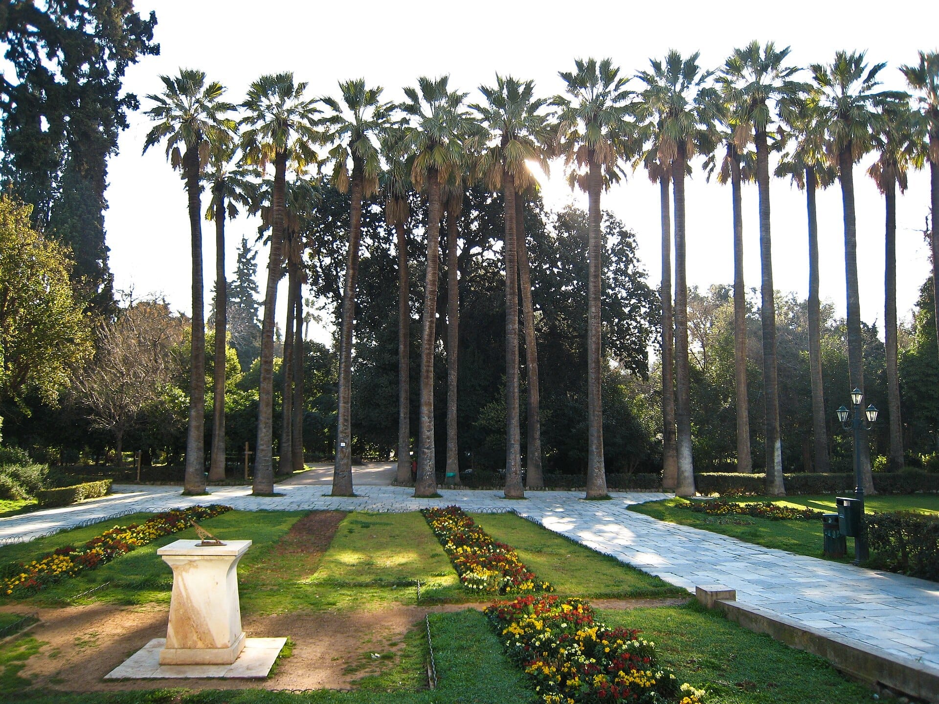 Alte palme, aiuole fiorite e una meridiana in pietra nel vialetto centrale del Giardino Nazionale di Atene sotto la luce del sole.