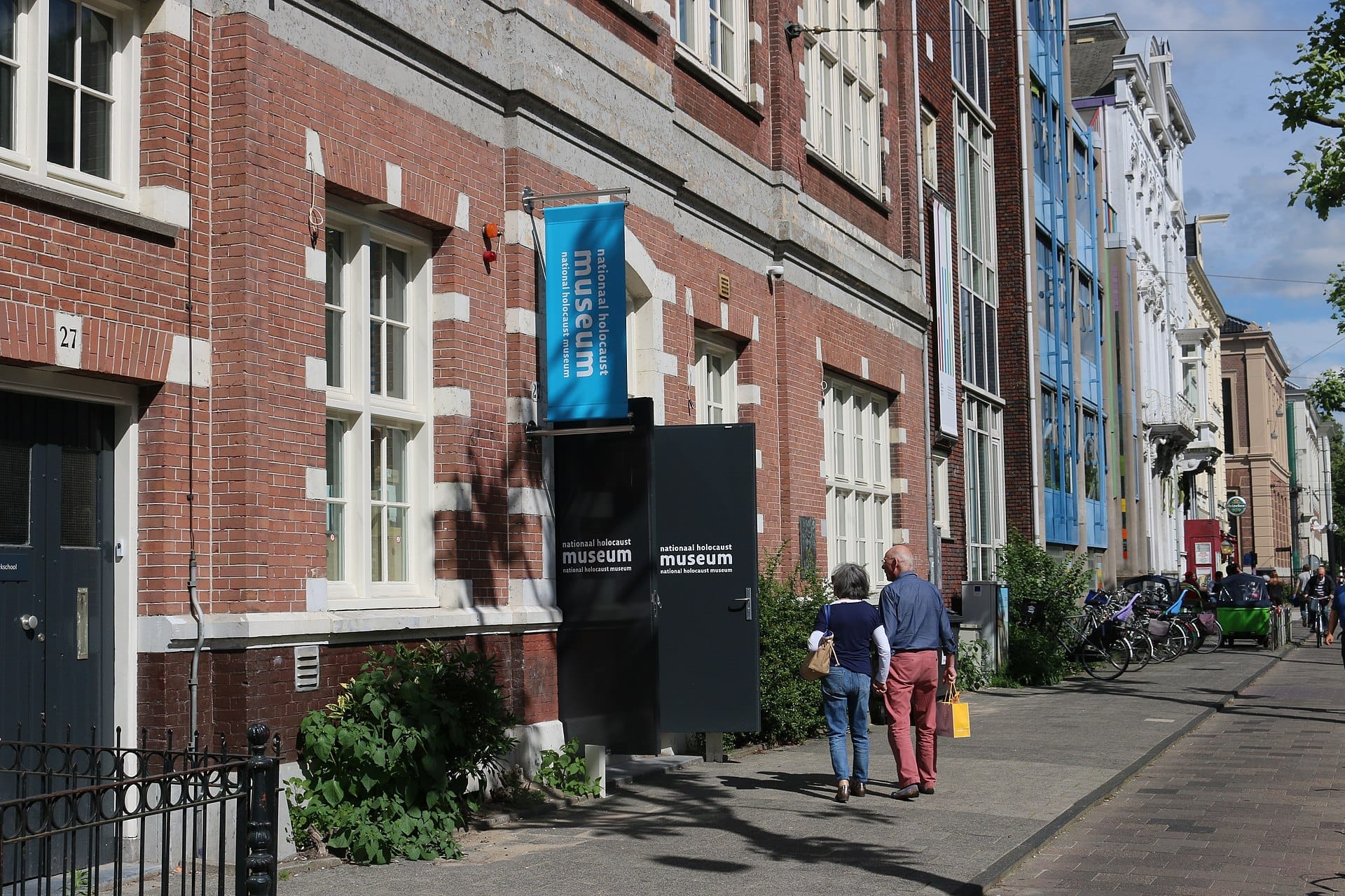 Vista exterior del National Holocaust Museum Amsterdam con su fachada de ladrillo rojo, señalización azul y dos personas caminando en un día soleado.