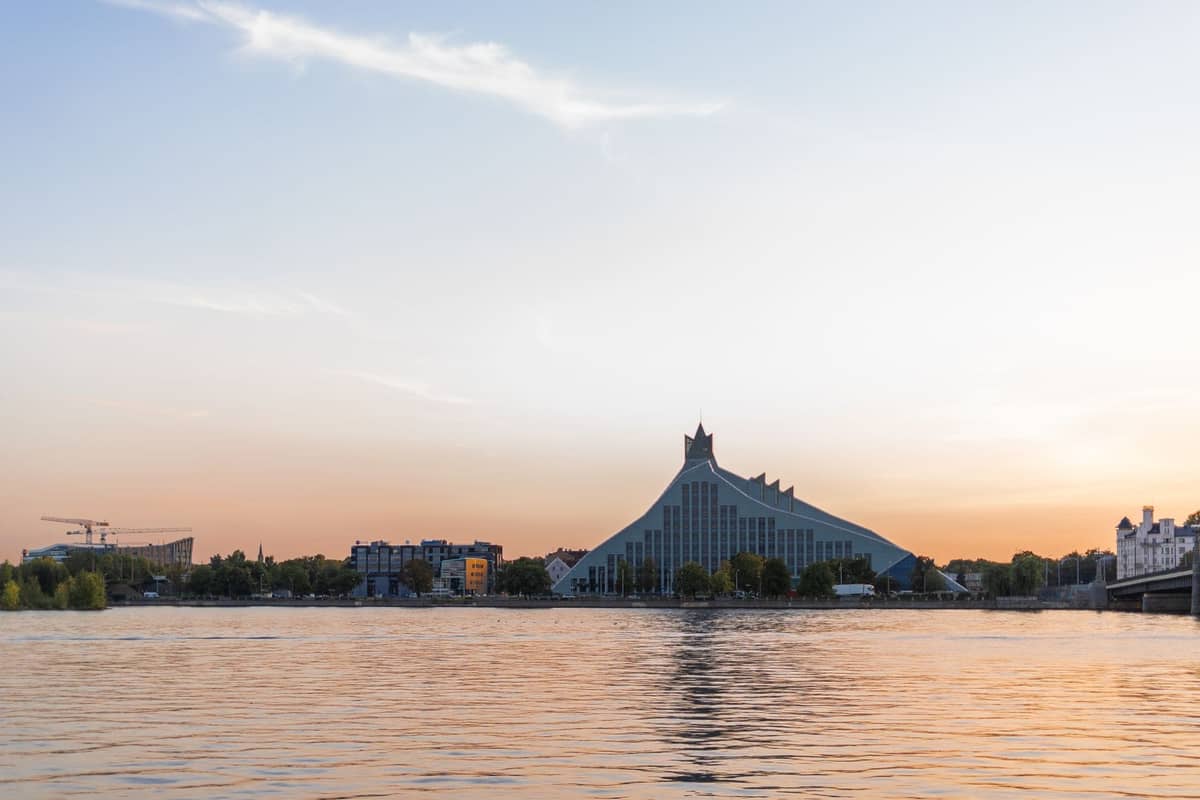 Die Lettische Nationalbibliothek mit ihrer markanten modernen Glasarchitektur, bei Sonnenuntergang von der anderen Seite der Daugava aufgenommen, der Himmel in sanften Farben leuchtend.