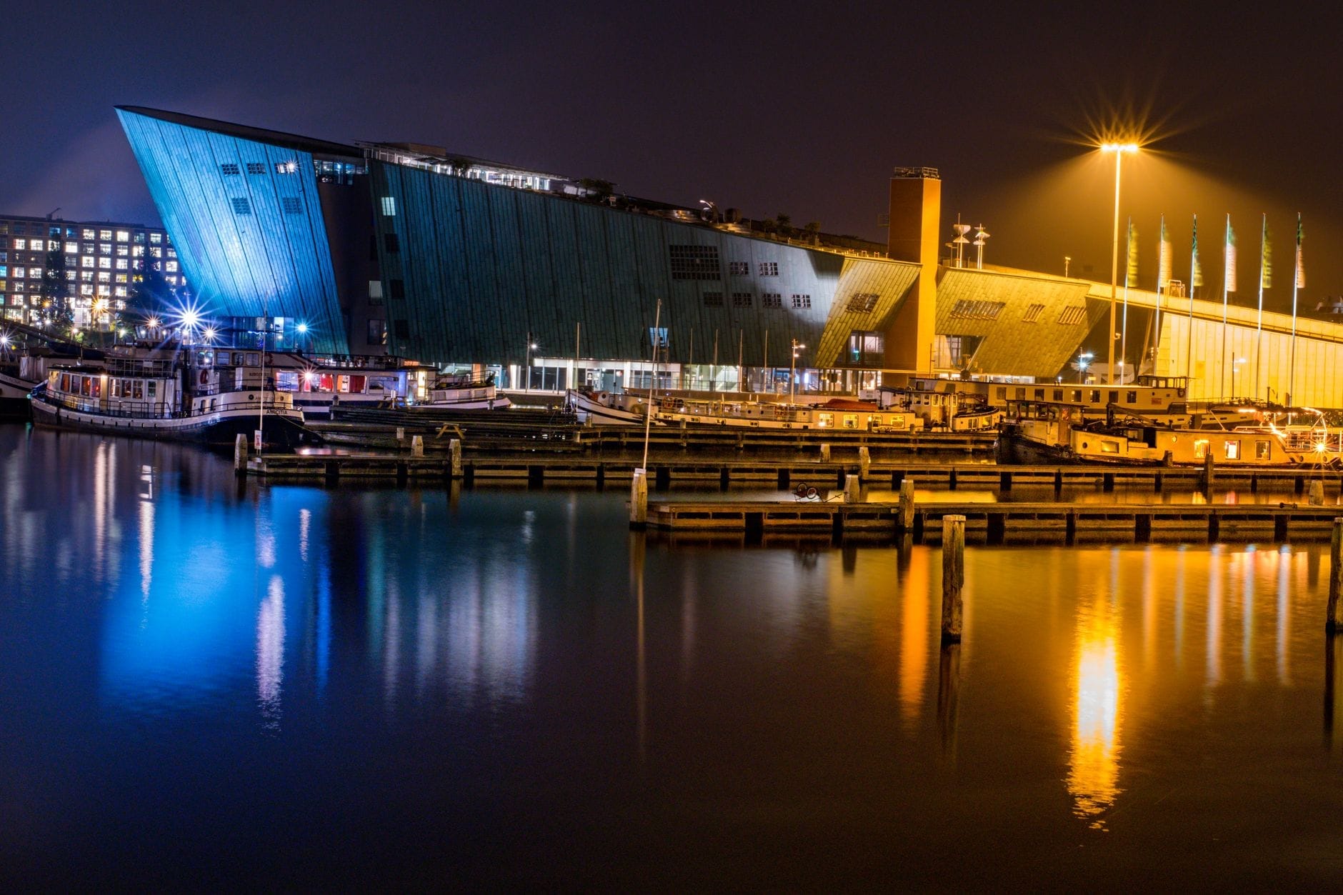 Vista nocturna de un museo marítimo moderno frente al agua, con una arquitectura llamativa que recuerda a un barco y cuyas luces se reflejan en las tranquilas aguas del puerto de Ámsterdam.