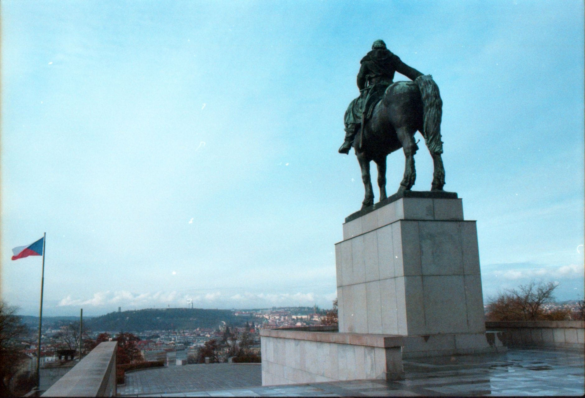 Reiterstatue auf dem Nationaldenkmal am Vítkov-Hügel mit Blick auf Prag, tschechischer Flagge und Stadtsilhouette unter blauem Himmel.