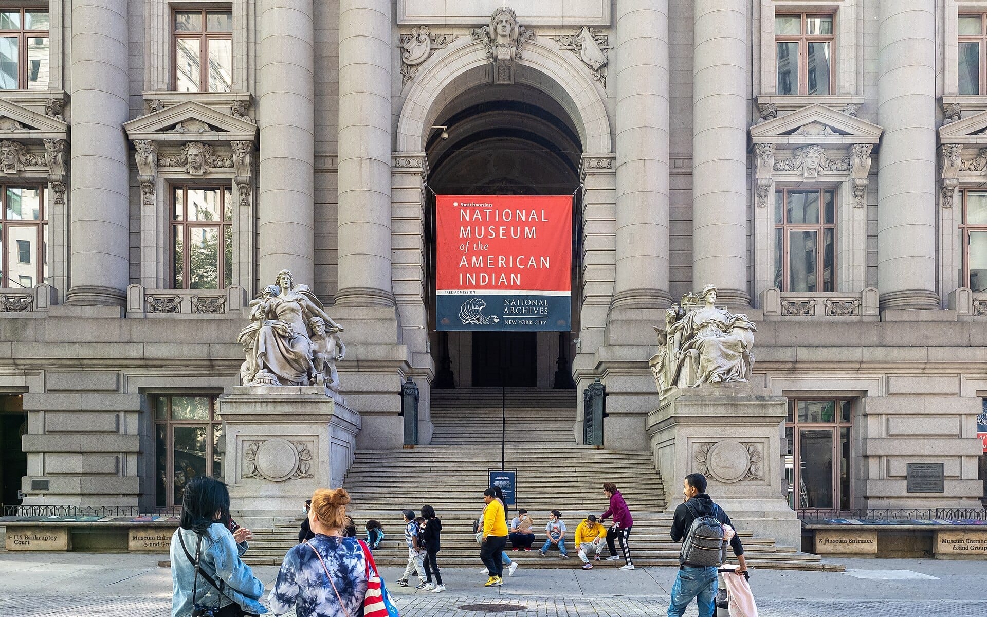 Il grande ingresso del National Museum of the American Indian presenta colonne massicce, statue elaborate e visitatori riuniti all'esterno in una giornata di sole.