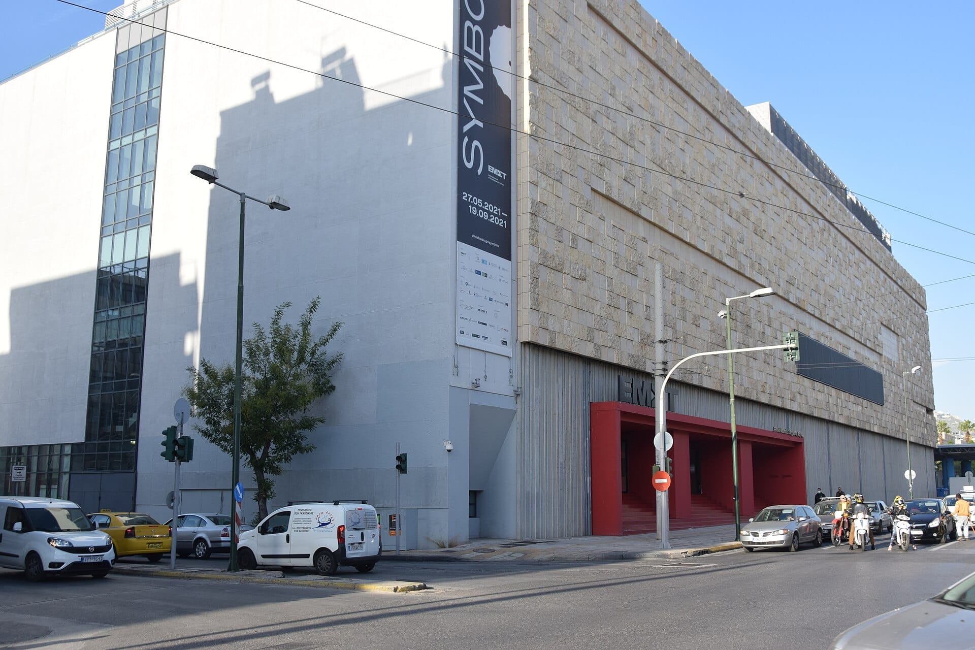 Vista della strada dell'edificio dell'EMST Atene con esterno in pietra e cemento, auto parcheggiate lungo la strada e un grande striscione verticale del museo.