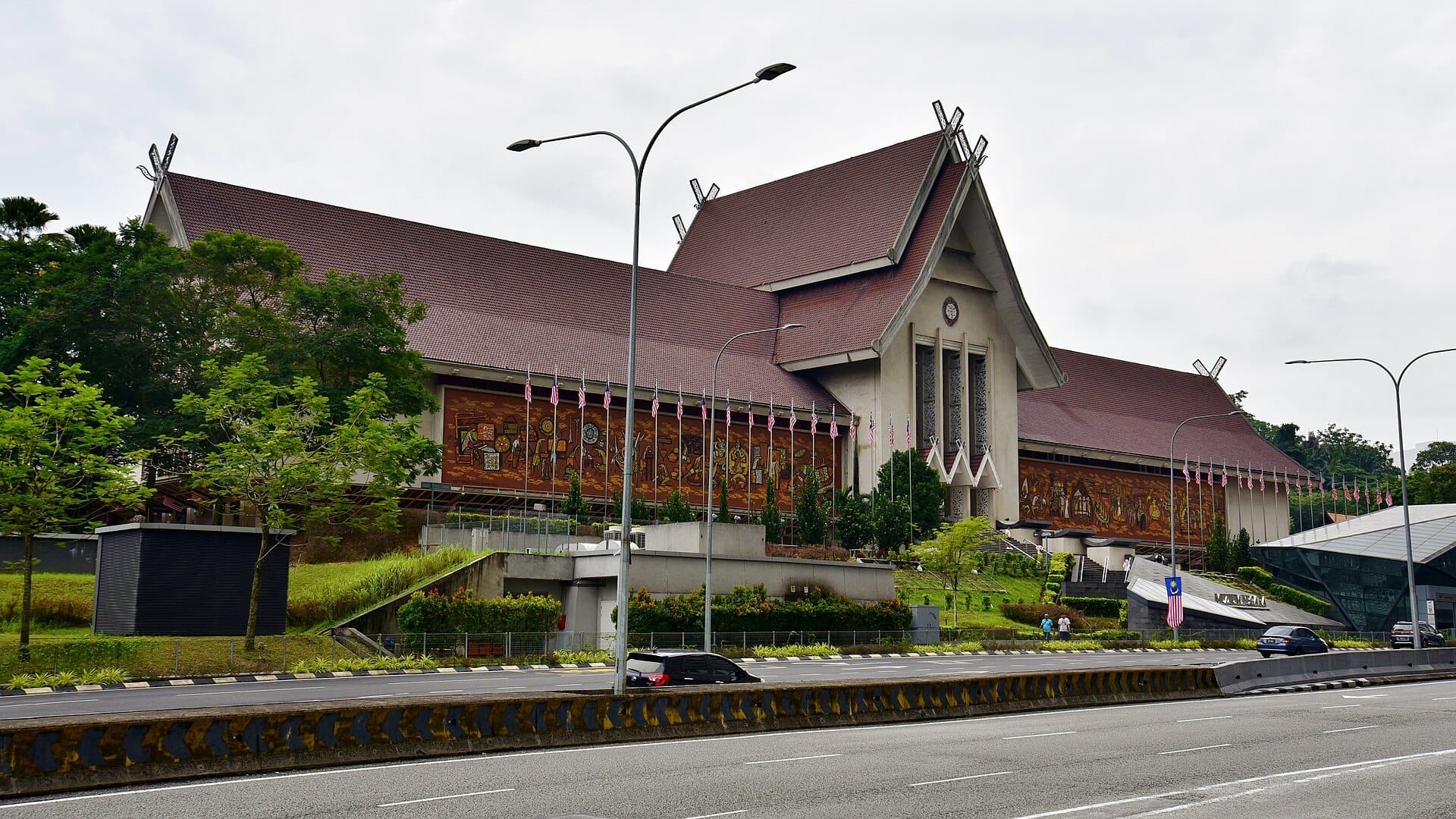 National Museum Malaysia exterior with traditional sloped roof, vibrant murals, and Malaysian flags lining the building on a cloudy day in Kuala Lumpur.