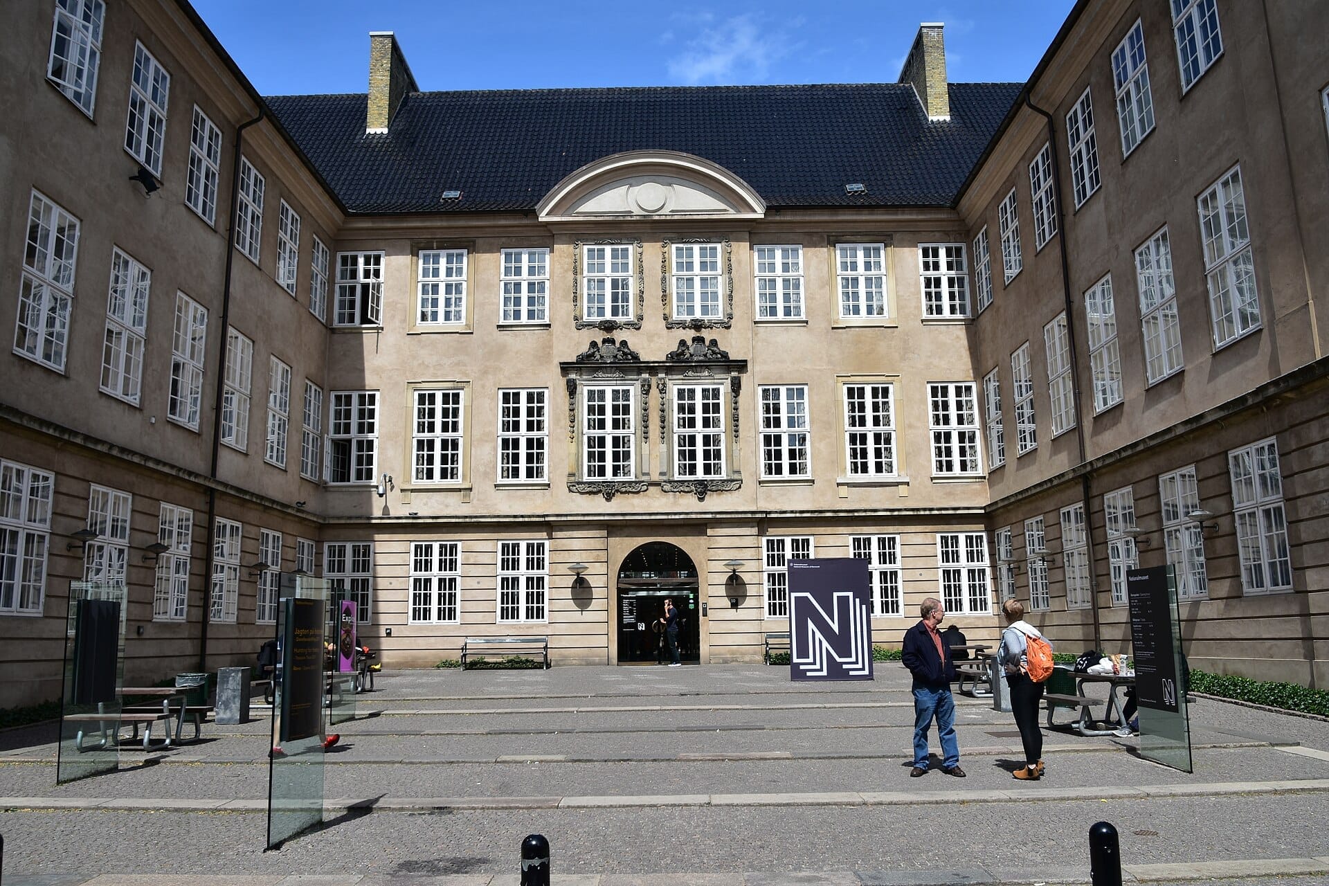 Cortile del Museo Nazionale della Danimarca con facciata storica, grandi finestre e visitatori vicino a moderni pannelli informativi in una giornata di sole.