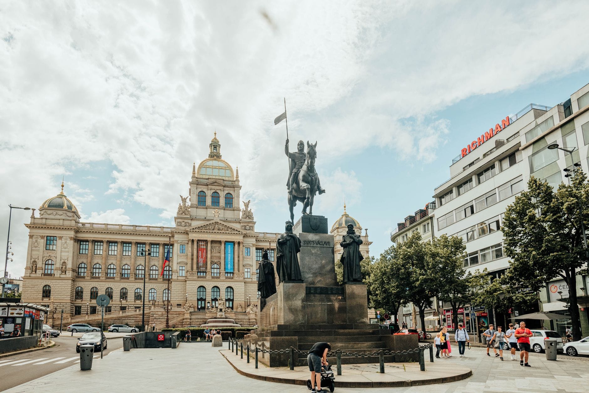 A Praça Venceslau com o Museu Nacional de Praga ao fundo, animada com pessoas, estátua, árvores e céu azul de verão, criando uma atmosfera de viagem convidativa.