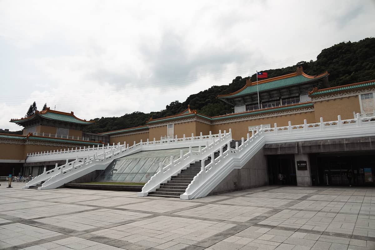 Weitwinkelaufnahme der Außenansicht des Nationalpalastmuseums in Taipei, Taiwan, mit der großen Eingangsfreitreppe, weißen Balustraden und grün bedachten Gebäuden vor einem bewaldeten Hang.