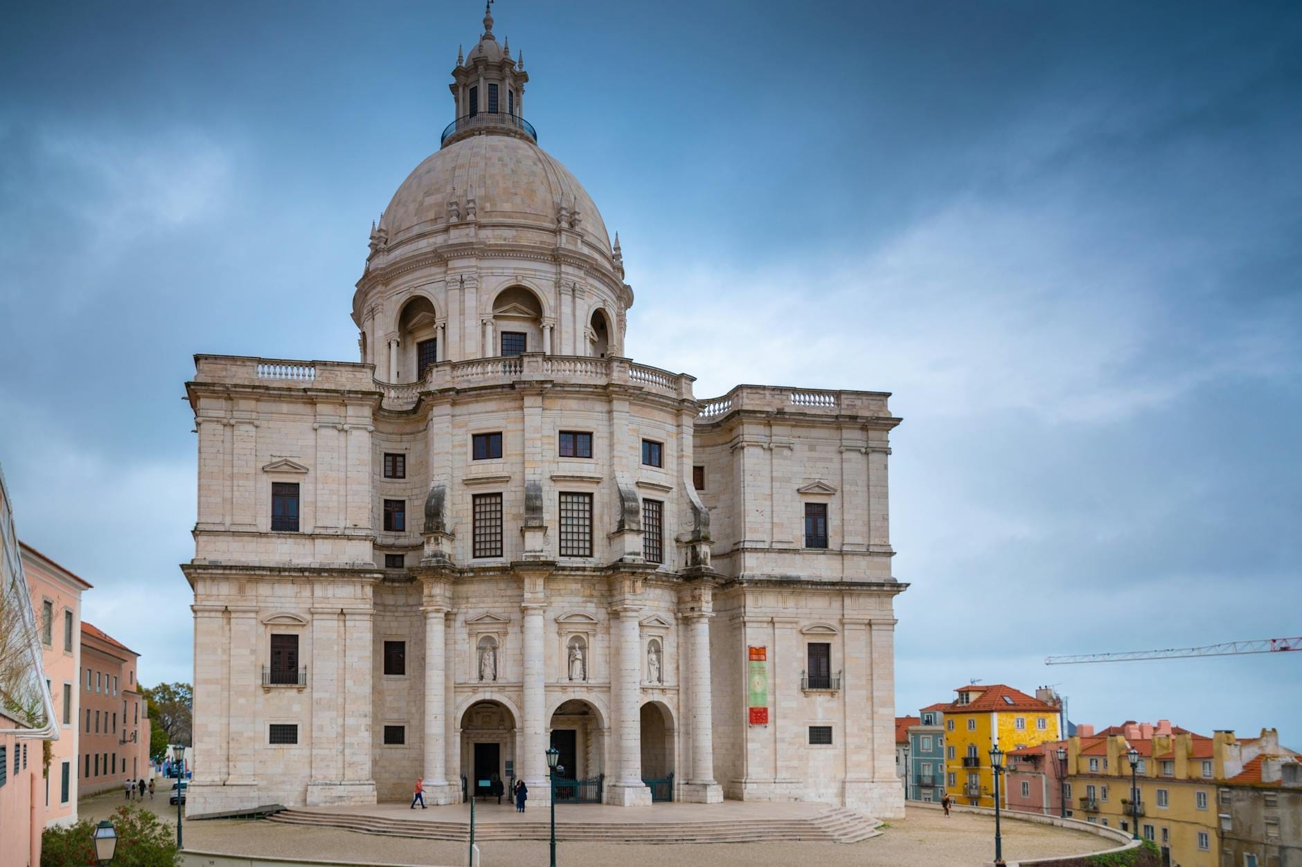 Vista frontale del Pantheon Nazionale di Lisbona sotto un cielo drammatico, con la facciata barocca e la cupola iconica sullo sfondo di case colorate.