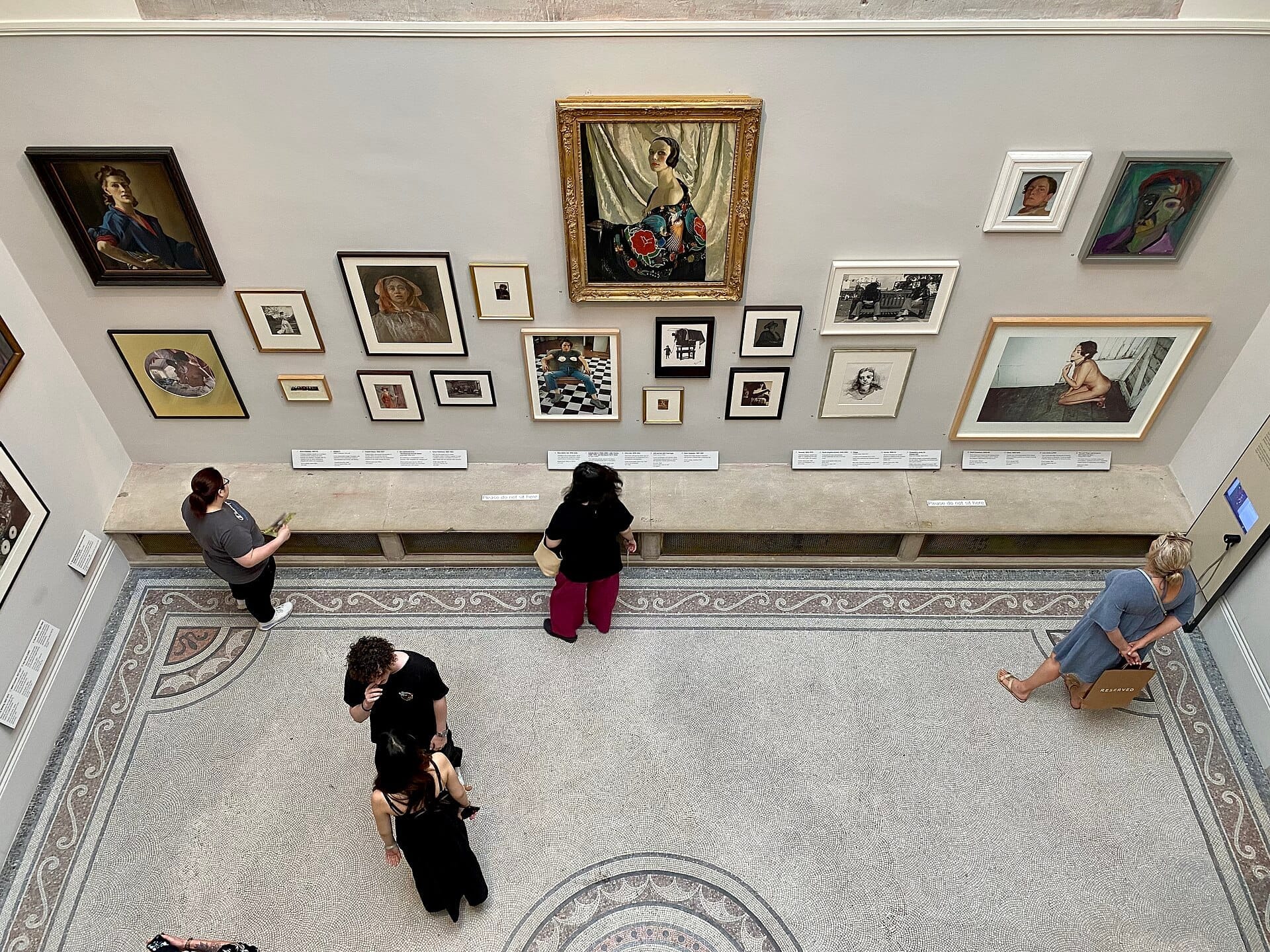 Visitatori che osservano un'eclettica collezione di ritratti incorniciati esposti su una parete di colore chiaro all'interno della National Portrait Gallery di Londra.