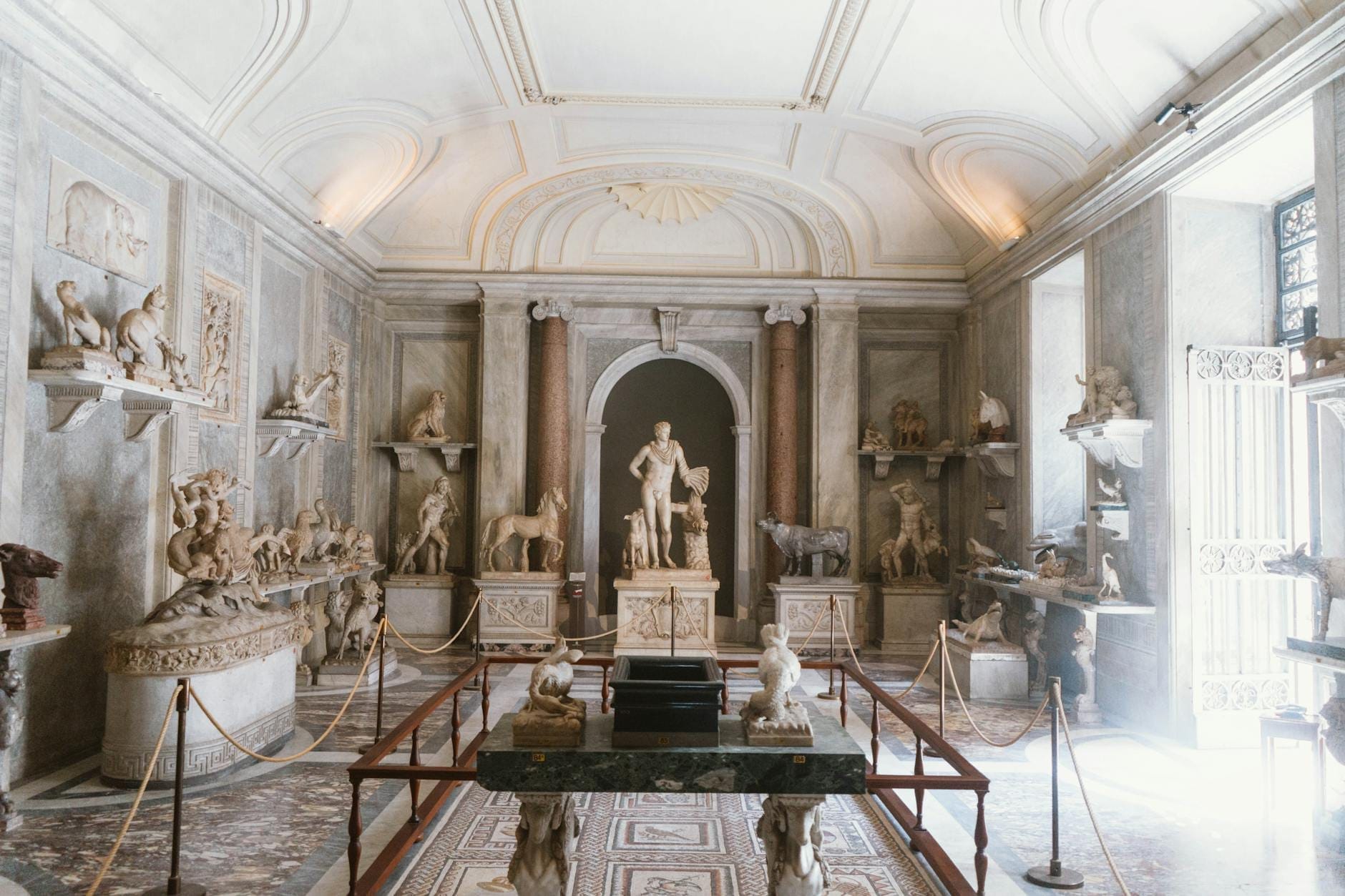 Elegant hall in a Roman museum filled with classical statues and marble sculptures, under a high arched ceiling with natural light streaming in.