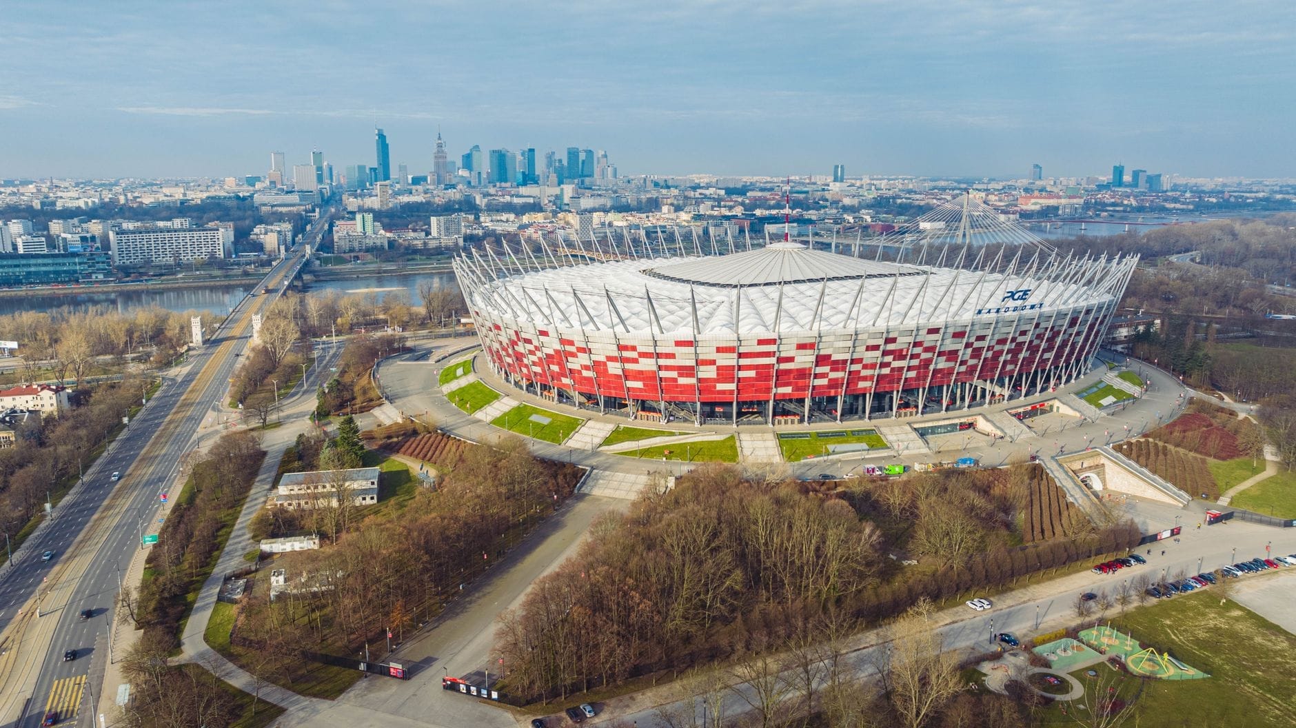ภาพมุมสูงของสนามกีฬา PGE Narodowy ริมฝั่งตะวันออกแม่น้ำวิสวา เห็นเส้นขอบฟ้าวอร์ซอว์ ต้นไม้ สะพาน และถนนล้อมรอบอารีนาแดงขาวทันสมัย