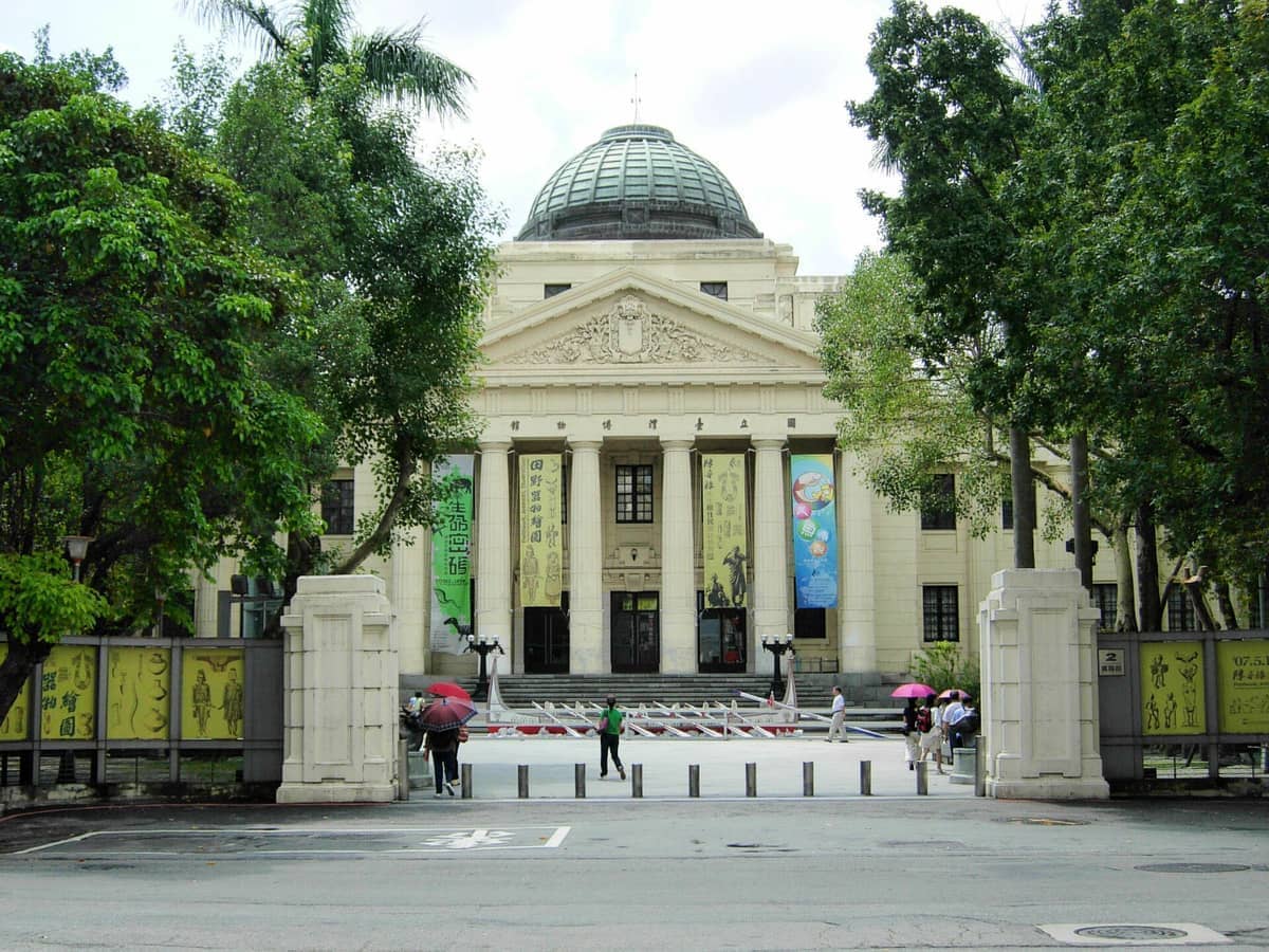 Frontansicht der prachtvollen Barockfassade des National Taiwan Museum, flankiert von hohen Bäumen und belebt von Besuchern mit Regenschirmen.