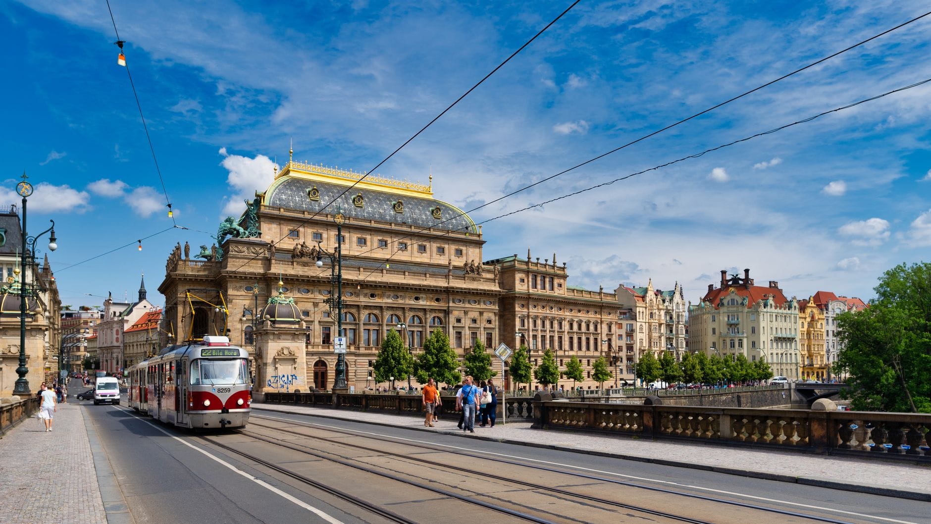 View of Prague's National Theatre with its golden roof, tram passing in front, blue sky, and lively street atmosphere along the Vltava river.