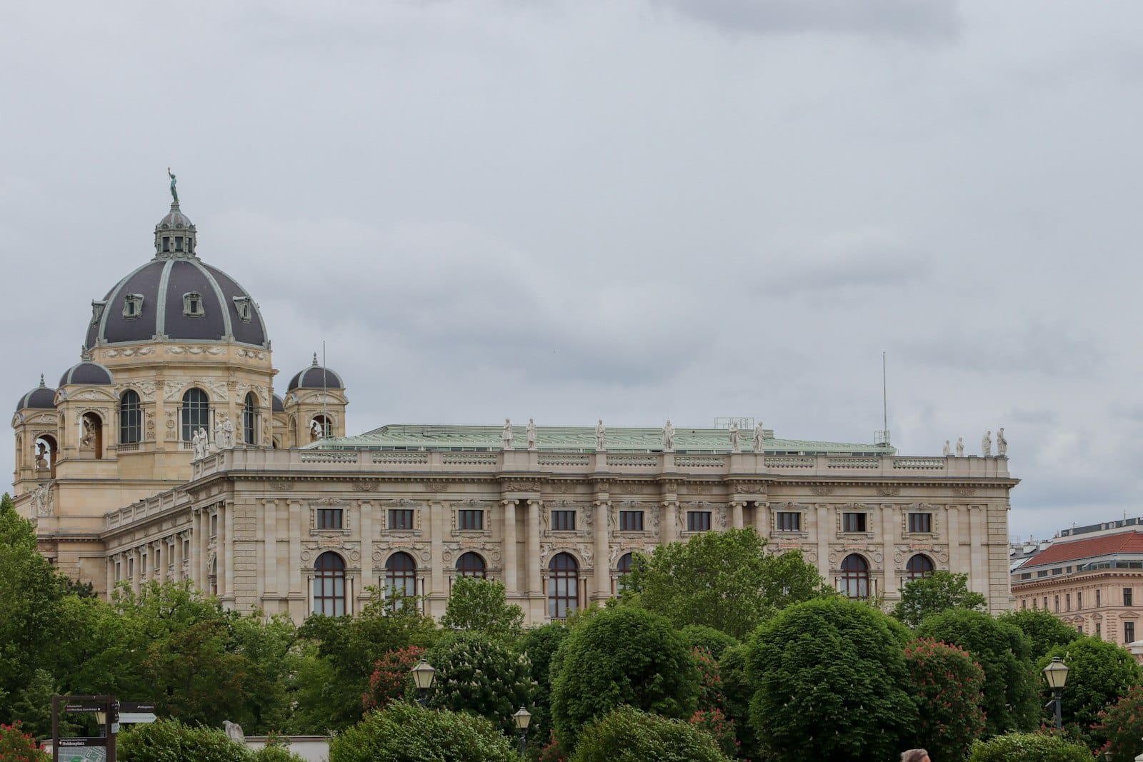 Weitwinkelperspektive des Naturhistorischen Museums Wien mit seiner beeindruckenden Neorenaissance-Fassade und der Kuppel, die sich über üppiges Grün unter bewölktem Himmel erhebt.